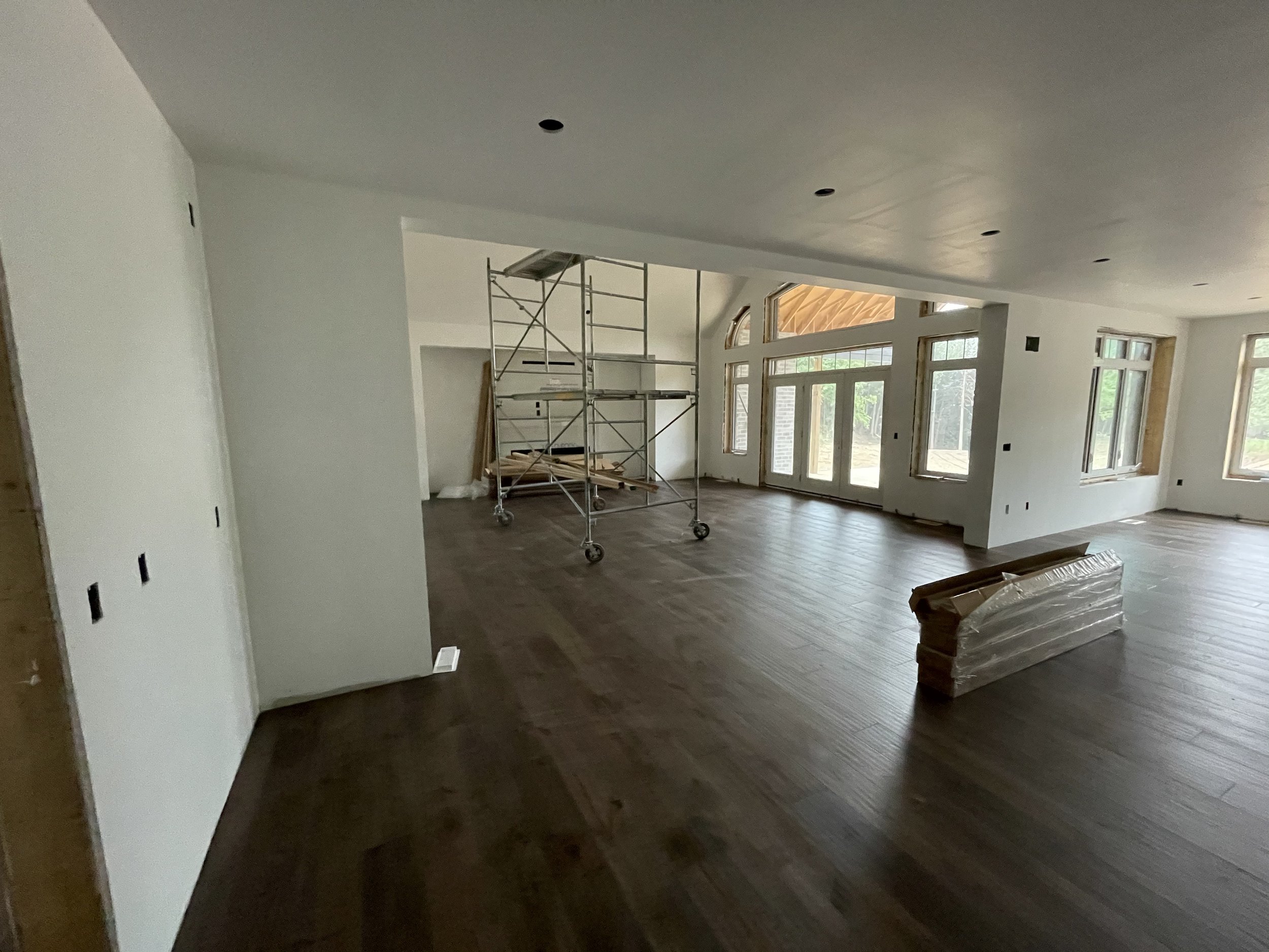 Interior of a house under construction or renovation, with white walls, large windows, and a wooden floor. Scaffolding is set up inside, and some construction materials are on the floor.