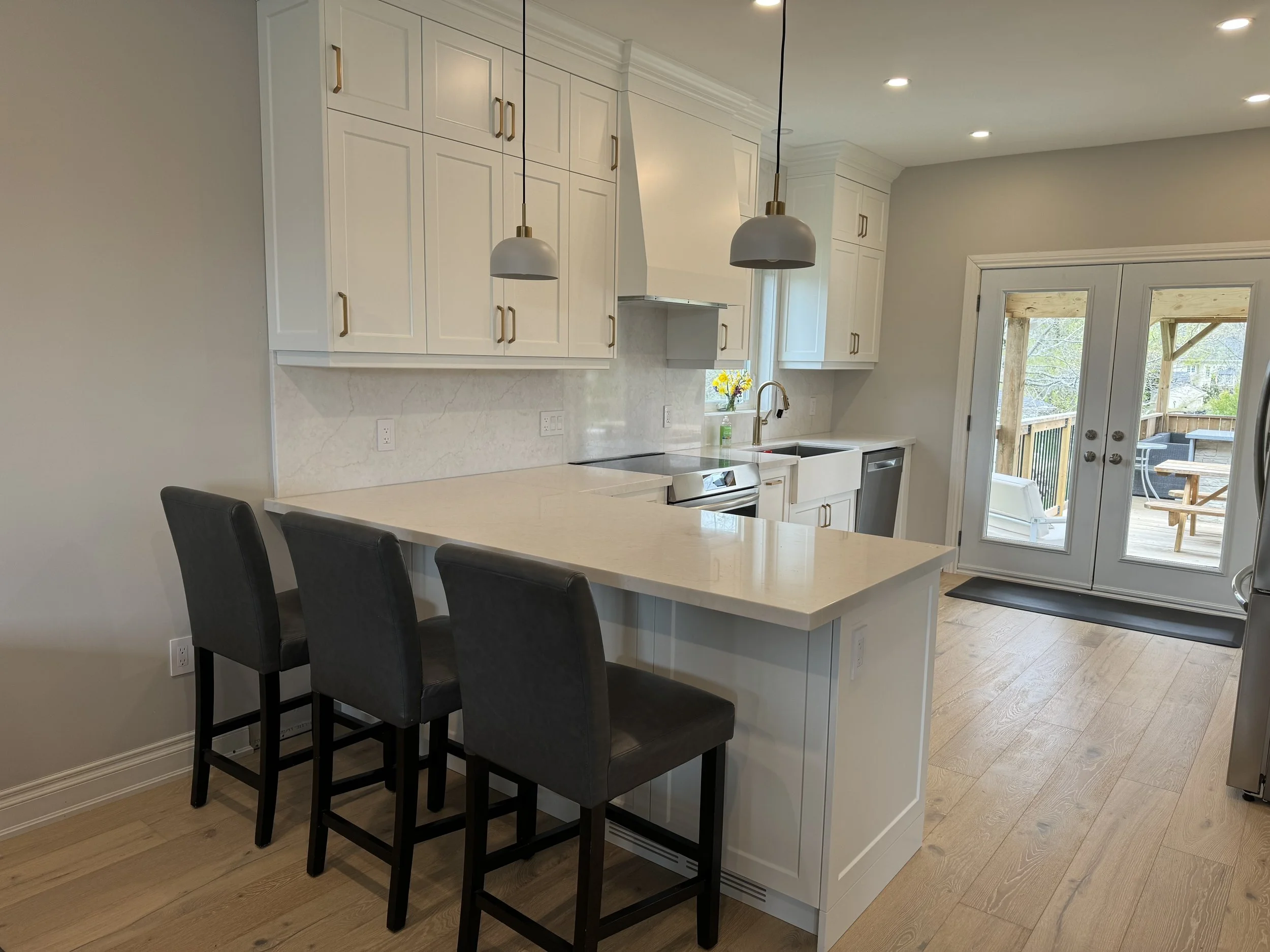 Modern Wasaga kitchen with white cabinets, marble backsplash, and an island with three black barstools. There are pendant lights hanging over the island, a window above the kitchen sink, and a set of glass double doors leading outside to a deck.