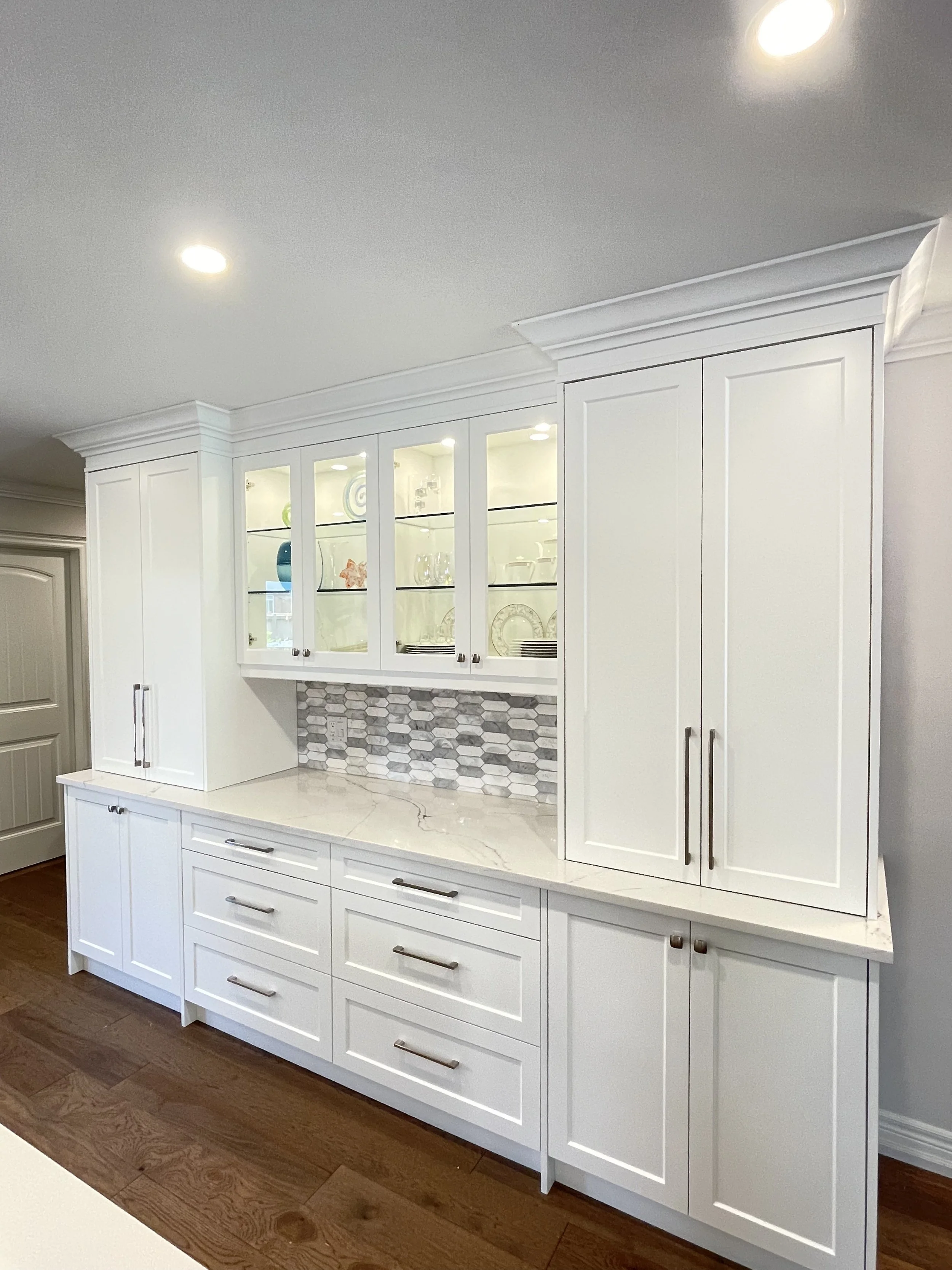 White kitchen cabinetry with glass-paneled upper cabinets displaying dishes and glassware, marble countertops, a gray and white hexagonal tile backsplash, and wood flooring.
