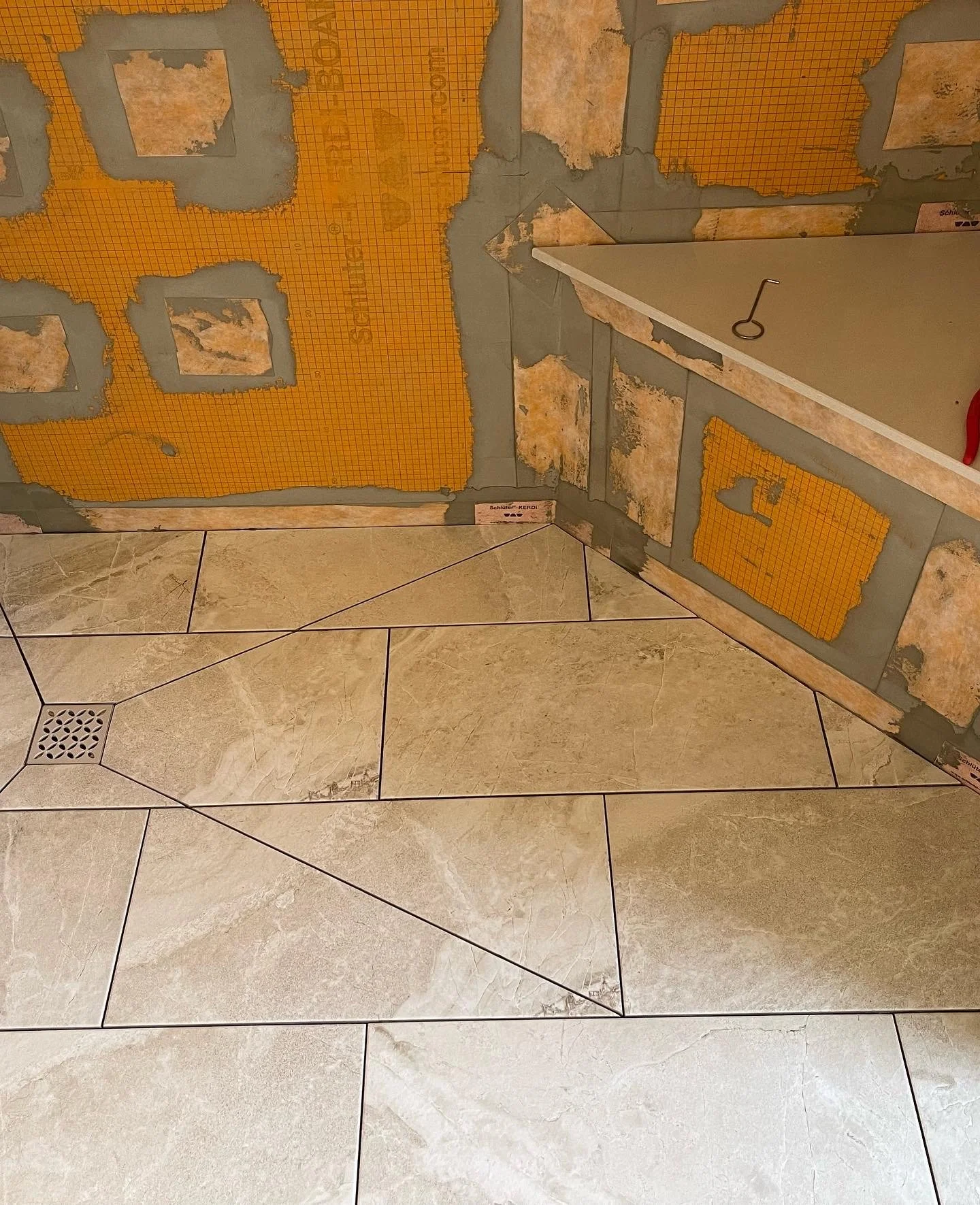 Partially tiled bathroom wall with Schluter membrane and exposed mortar, and beige tiled floor with a square drain cover.