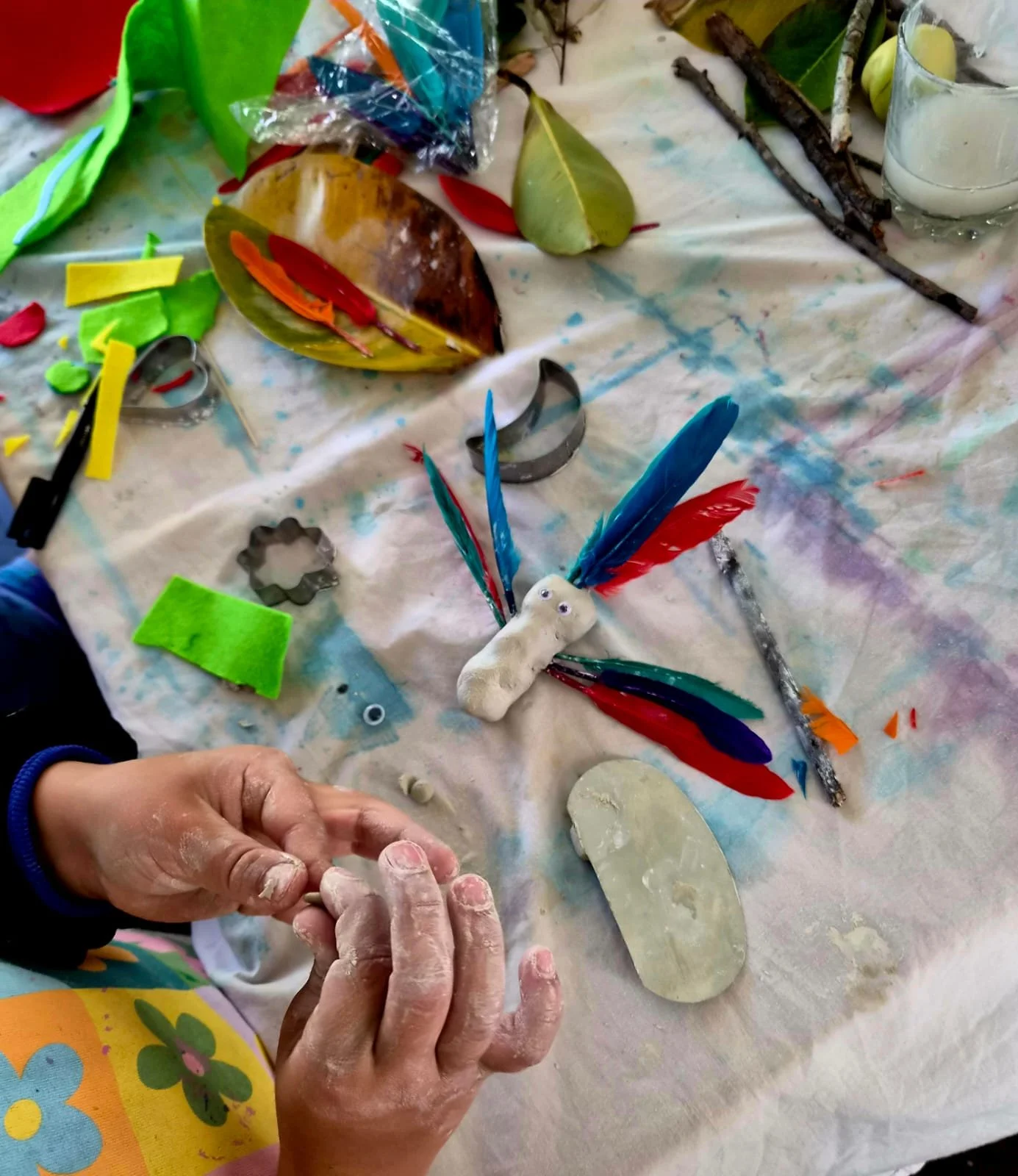 A person working on a craft project with colorful feathers, leaves, sticks, and various materials on a cloth-covered table. The person's hands are covered in clay or plaster, shaping a small object.