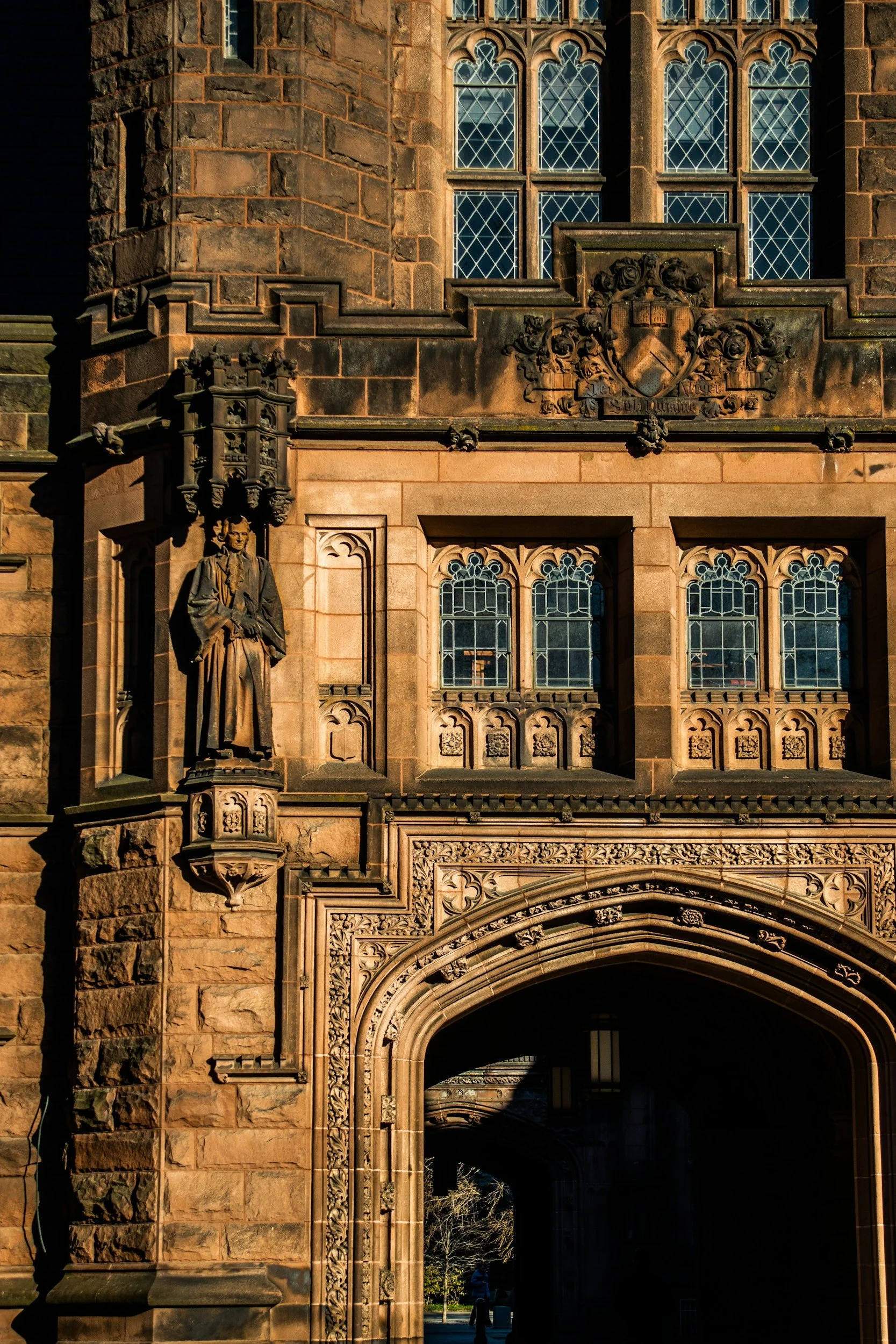 Close-up of an ornate stone building entrance with gothic-style windows, a sculpture of a historical figure, and detailed carvings on the stone facade, suggesting an old university or church.