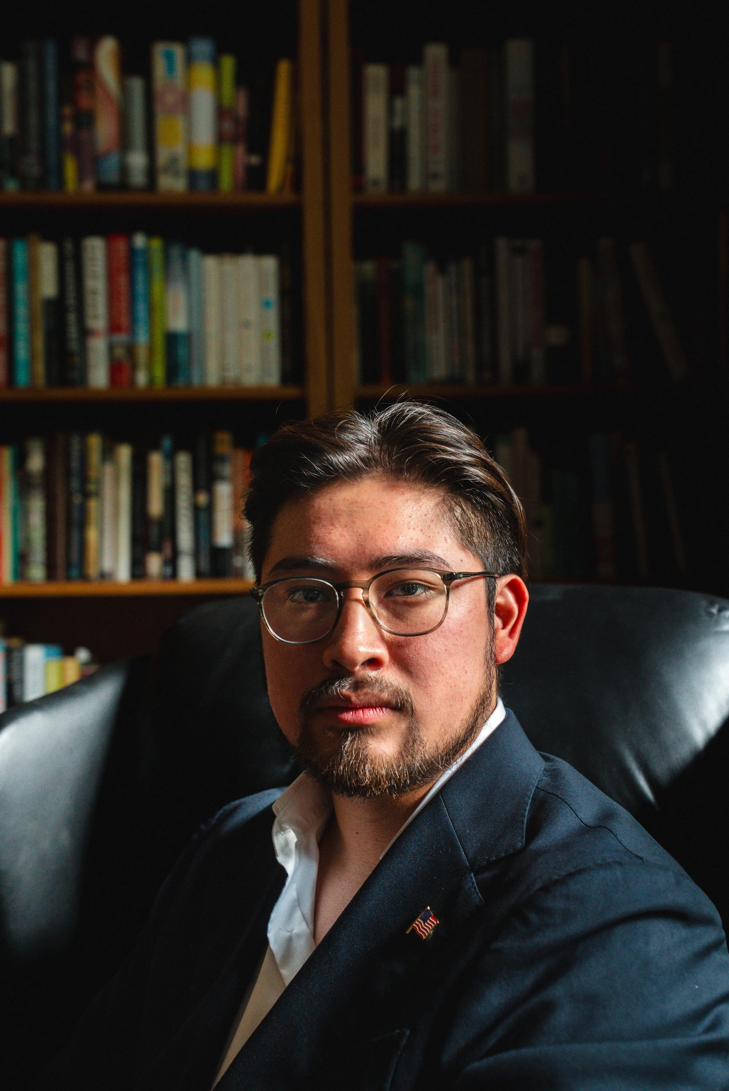 Portrait of a young man with glasses, dark hair, and a beard, wearing a suit and white shirt, sitting on a black leather chair in a room with bookshelves in the background.