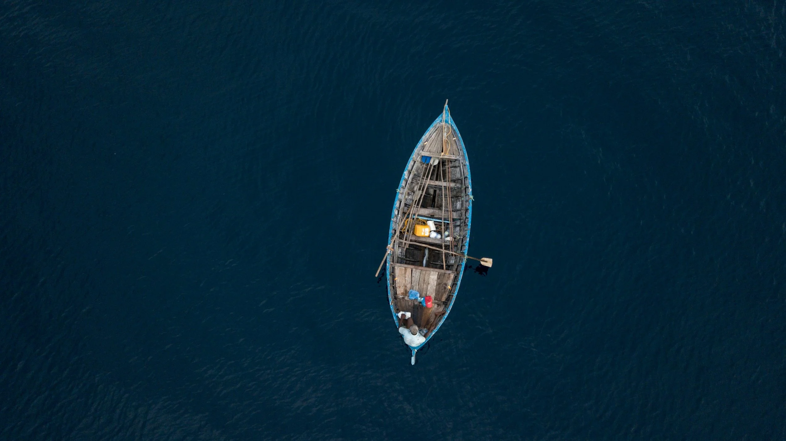 An aerial view of a small wooden boat floating in dark blue water, with various items inside including containers, a piece of fabric, and an oar.