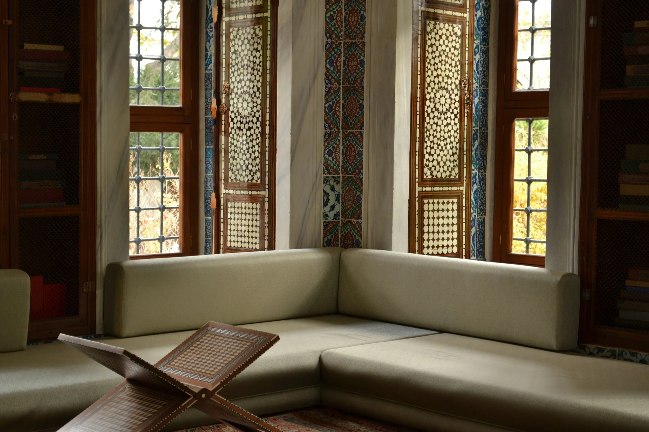 A corner of a room with large windows featuring decorative wooden frames and ornate tile inlays. A built-in bookshelf with books is visible on both sides of the windows. A cream-colored sectional sofa is placed in front, and there is a chessboard on a small foldable table.