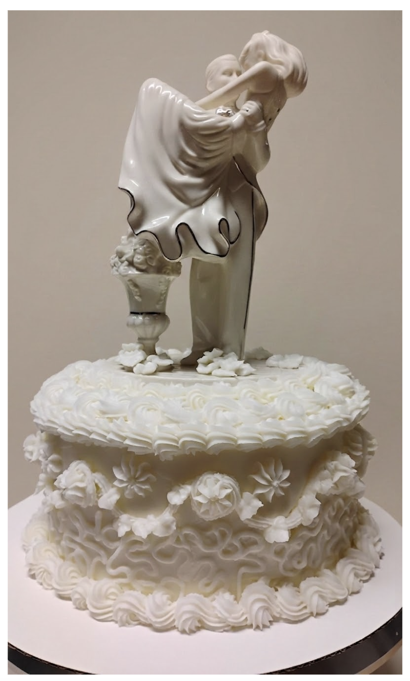 A white cake with floral and swirl decoration, topped with a sculpture of a woman and man embracing.