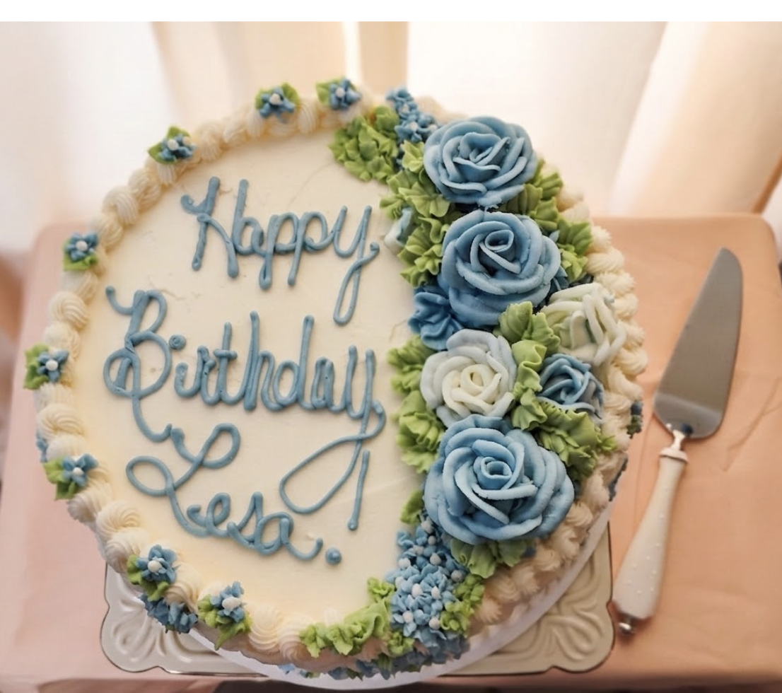 Round birthday cake with white icing, decorated with light blue, white, and green flowers on one side. The cake has a border  The message on the cake reads "Happy Birthday Le in blue cursive icing. A cake knife with a white handle and a silver blade 
