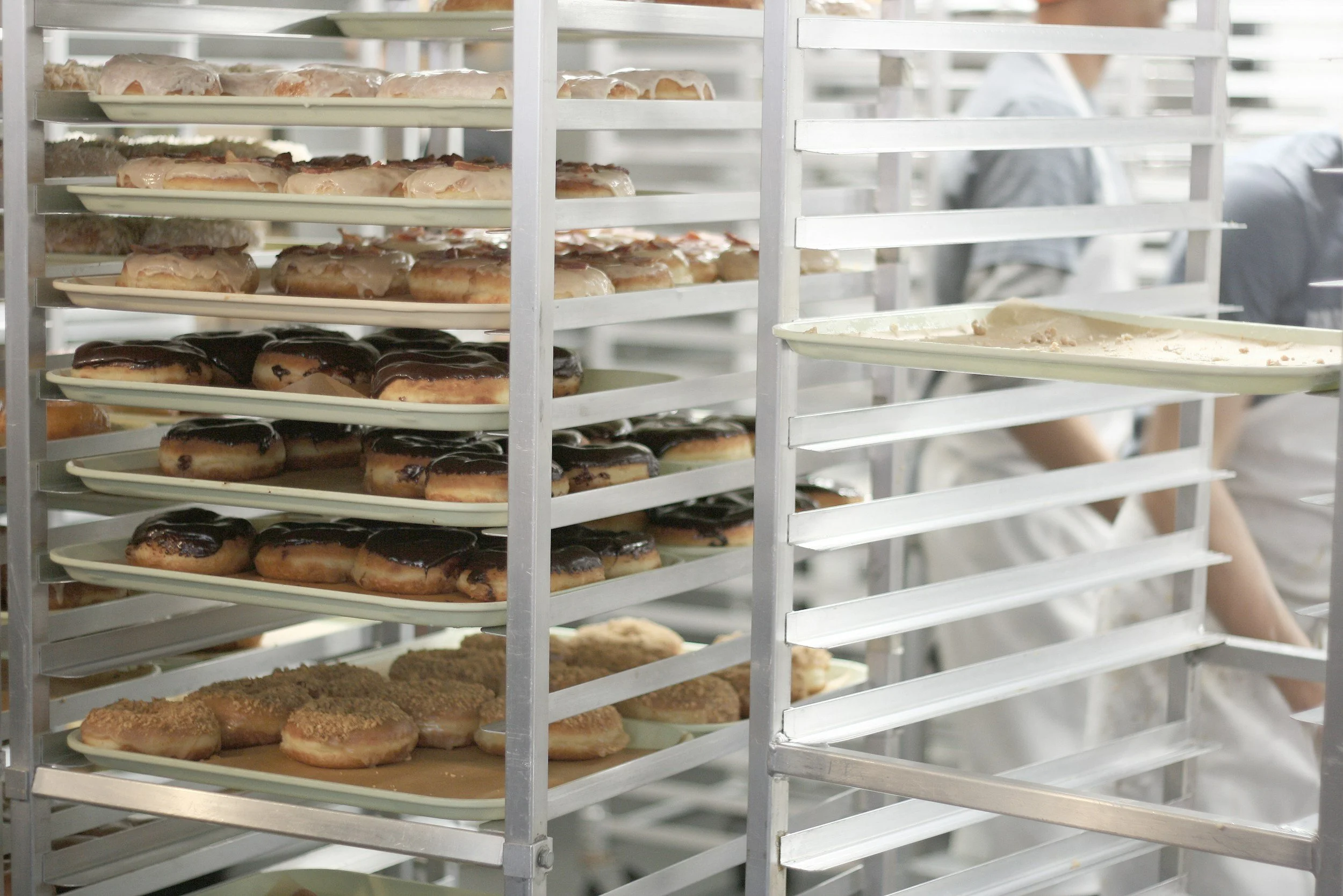 Trays of assorted glazed donuts, some with chocolate, others with pink or white glaze, on metal racks in a bakery or kitchen setting.