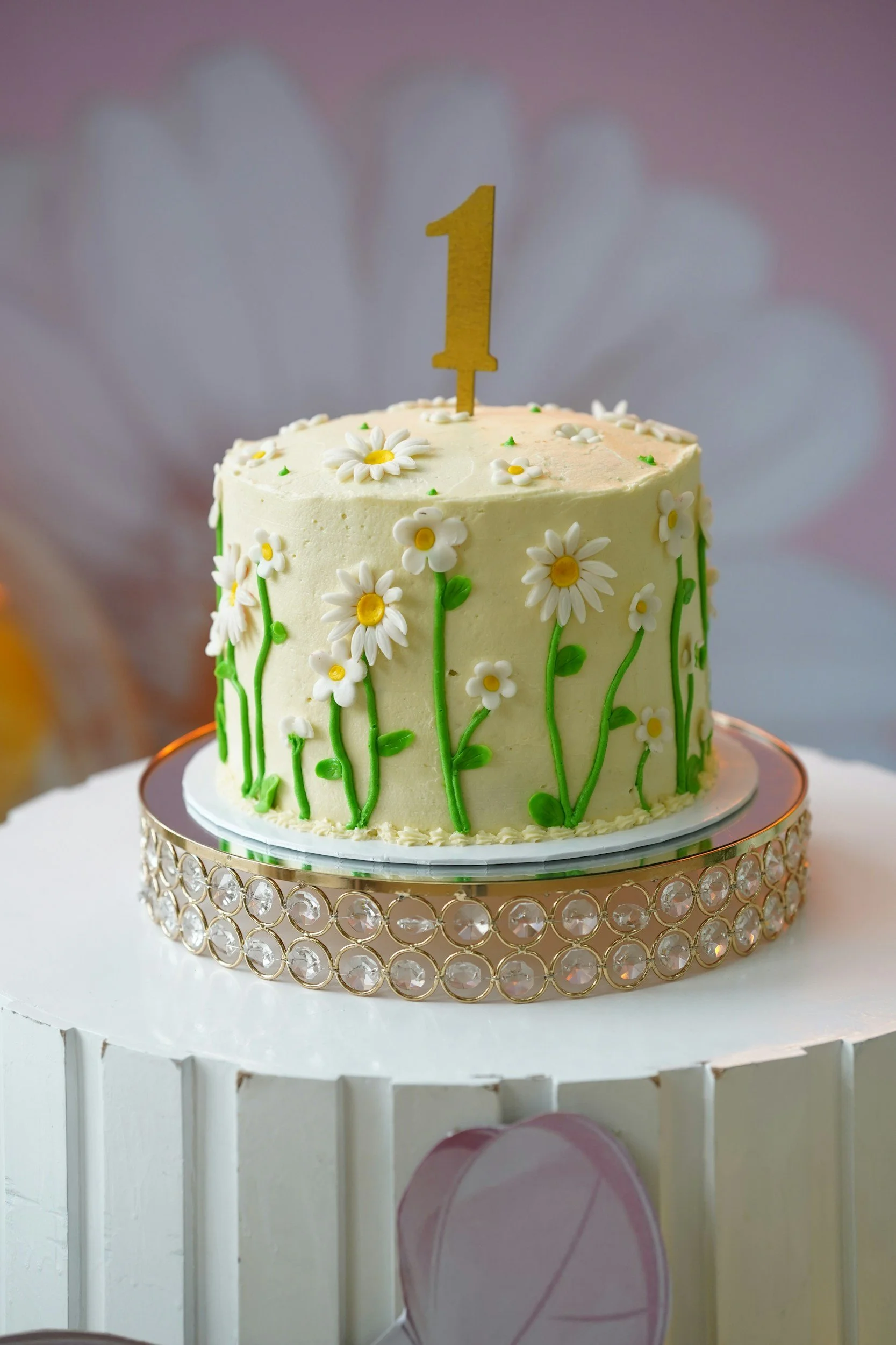A birthday cake decorated with white flowers and green stems, with a gold "1" cake topper, sitting on a round mirror atop a white table with a decorative crystal-like border.