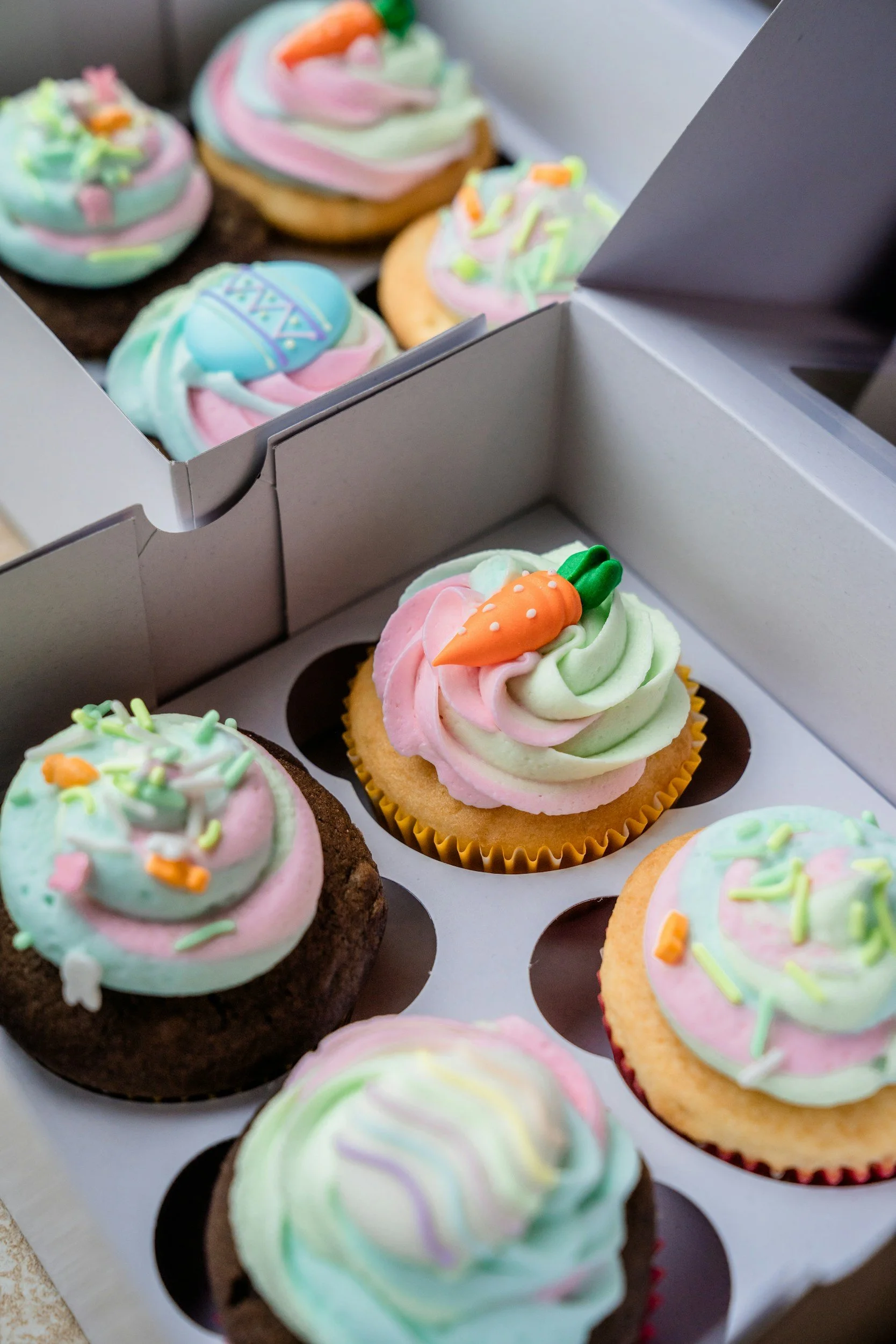 A box of decorated cupcakes with pastel-colored frosting and sprinkles, some with small sugar decorations, in a white bakery box.