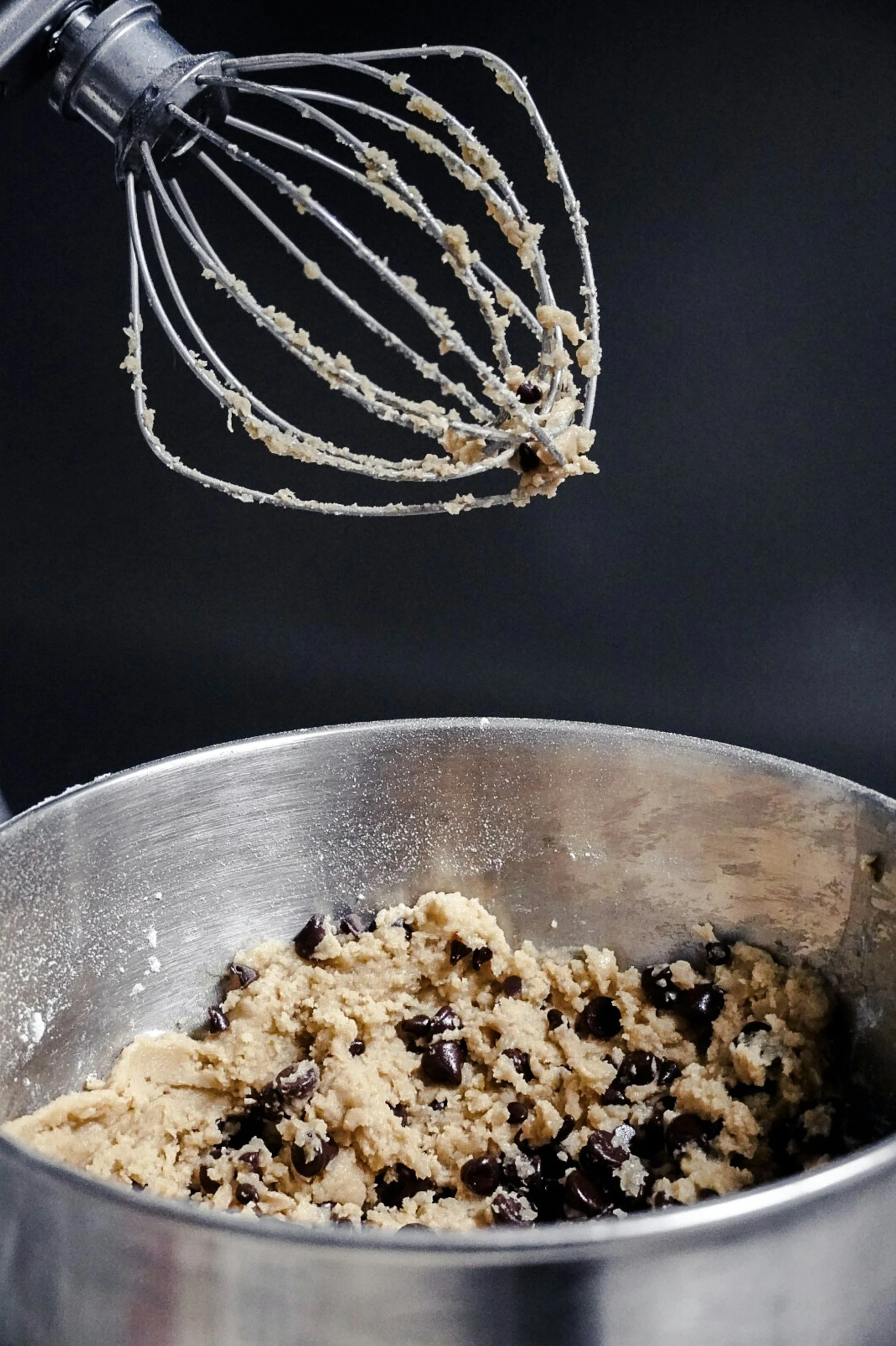 A metal stand mixer with cookie dough and chocolate chips inside a mixing bowl. The beater attachment is lifted above the bowl.
