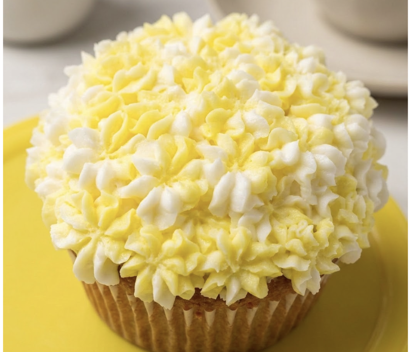 Cupcake topped with white and yellow frosting that looks like flower petals, on a yellow surface.