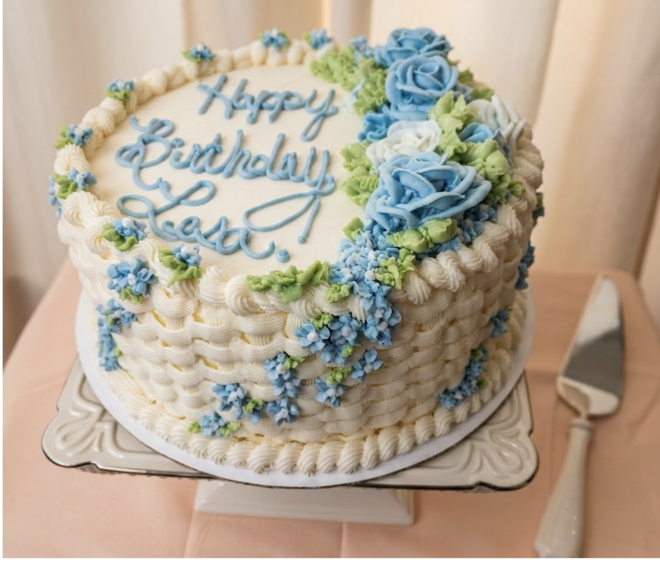 A round birthday cake decorated with white, blue, and green floral icing, with the words 'Happy Birthday Lesa written in blue icing on top. The cake is on a decorative cake stand, with a cake knife beside it.