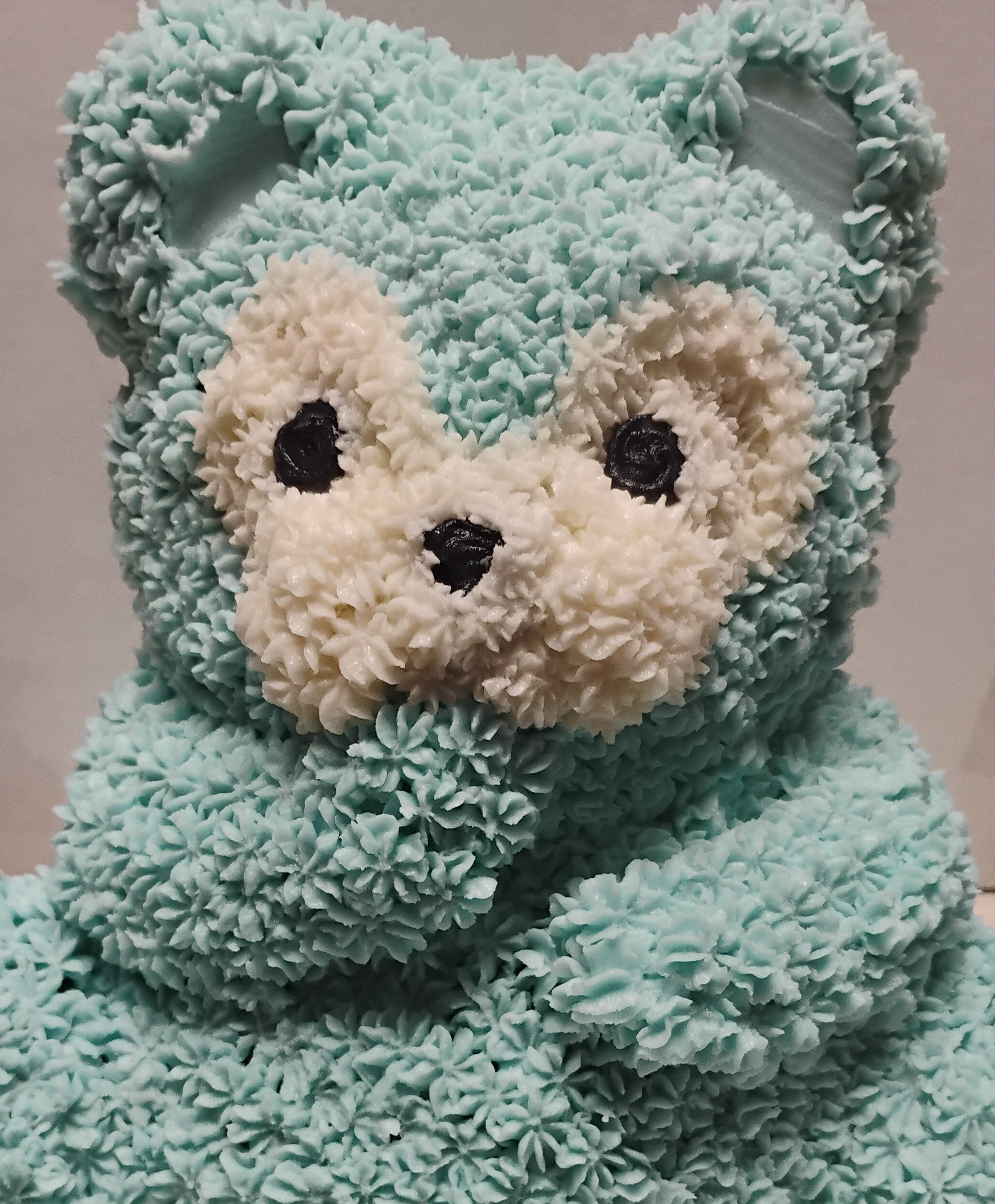 A cake designed to look like a cute, fluffy blue bear, with textured icing for fur, white icing for the face and ears, and black icing for the eyes and nose.