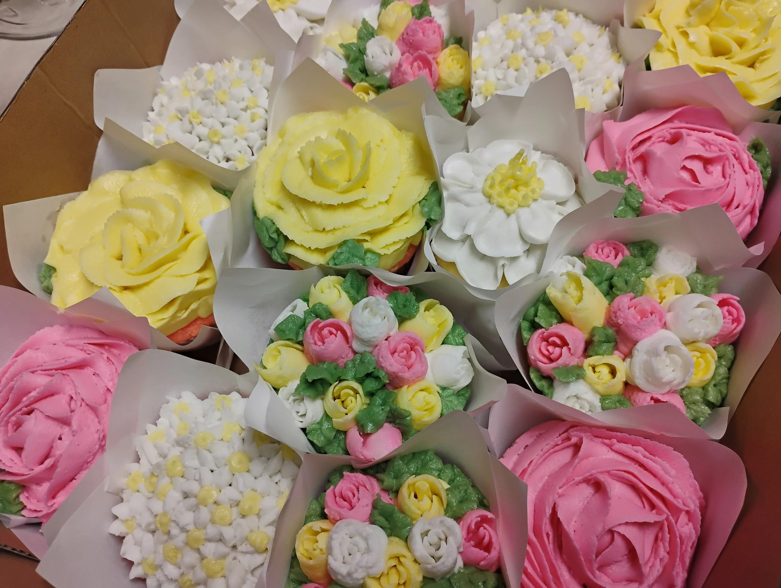 A collection of cupcakes decorated with various colorful floral-themed icing, including pink, white, yellow, and green designs resembling roses, daisies, and other flowers.