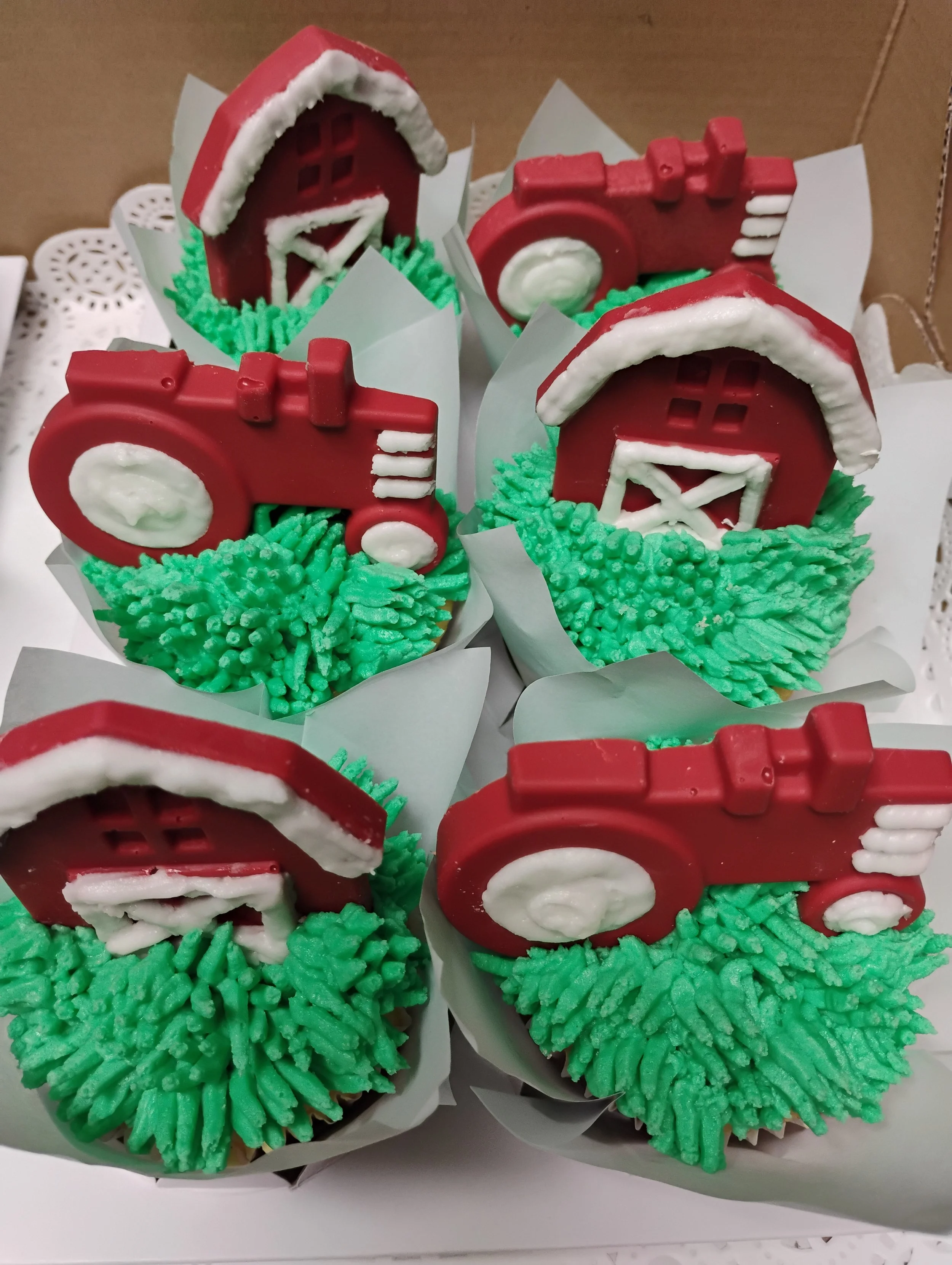 Decorative cupcakes shaped like red farm barns and red tractors, decorated with green frosting resembling grass.