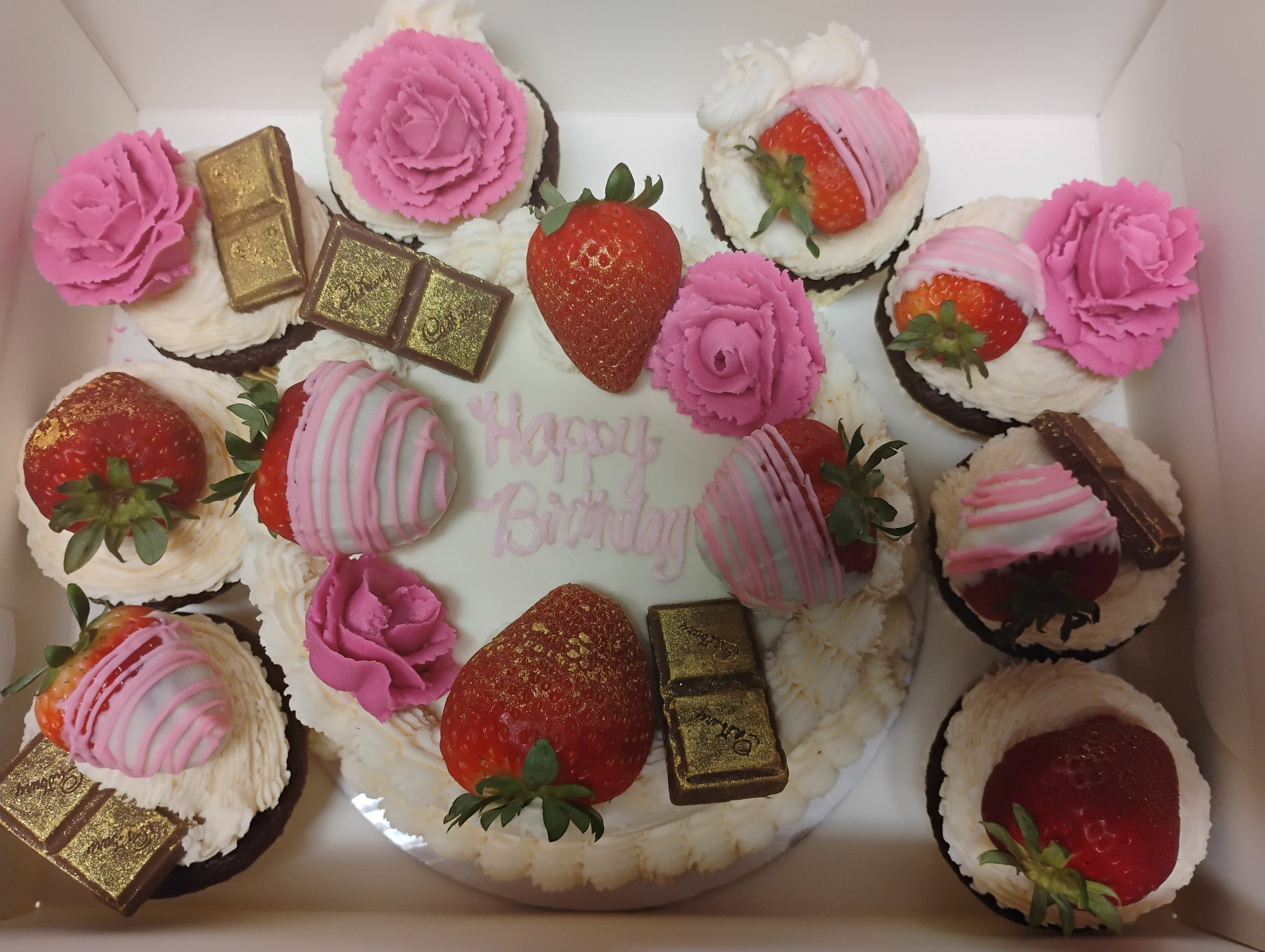A birthday cake topped with strawberries, pink roses, white and pink striped chocolate pieces, gold-dusted chocolates, and pink frosting flowers, with a message that says "Happy Birthday" in pink icing.
