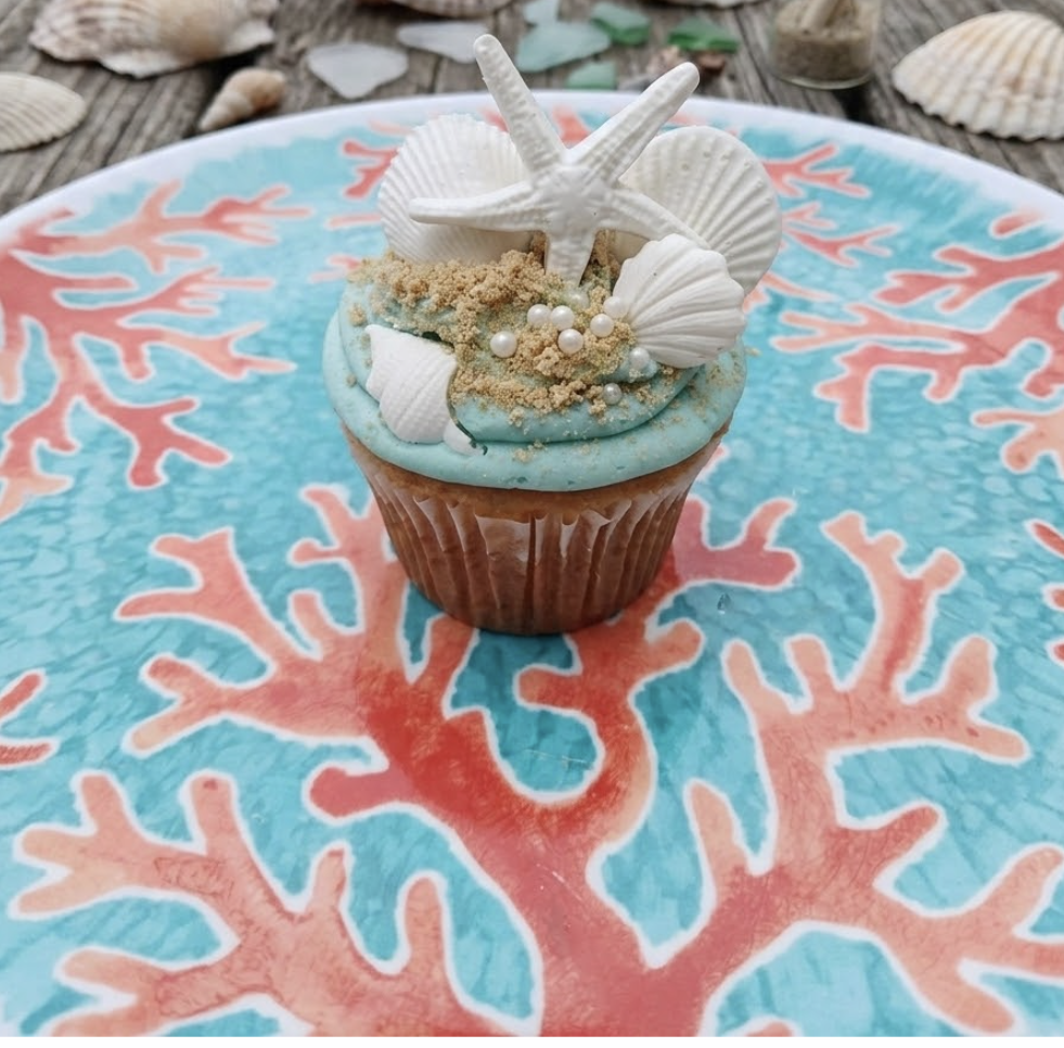 Cupcake with blue frosting decorated with white seashells, starfish, and edible pearls, placed on a colorful coral-patterned plate.