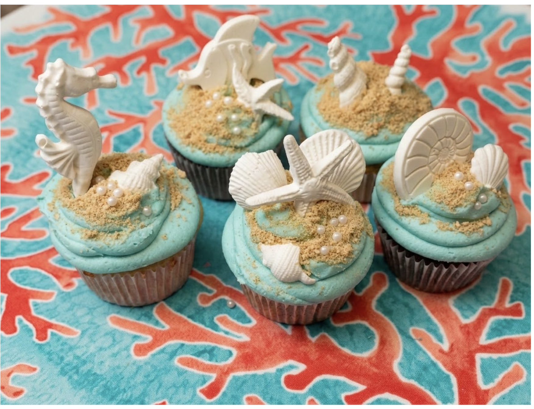 Six cupcakes with sea-themed decorations, including white seashell, starfish, seahorse, and seashell toppers, on a colorful coral-patterned cloth.