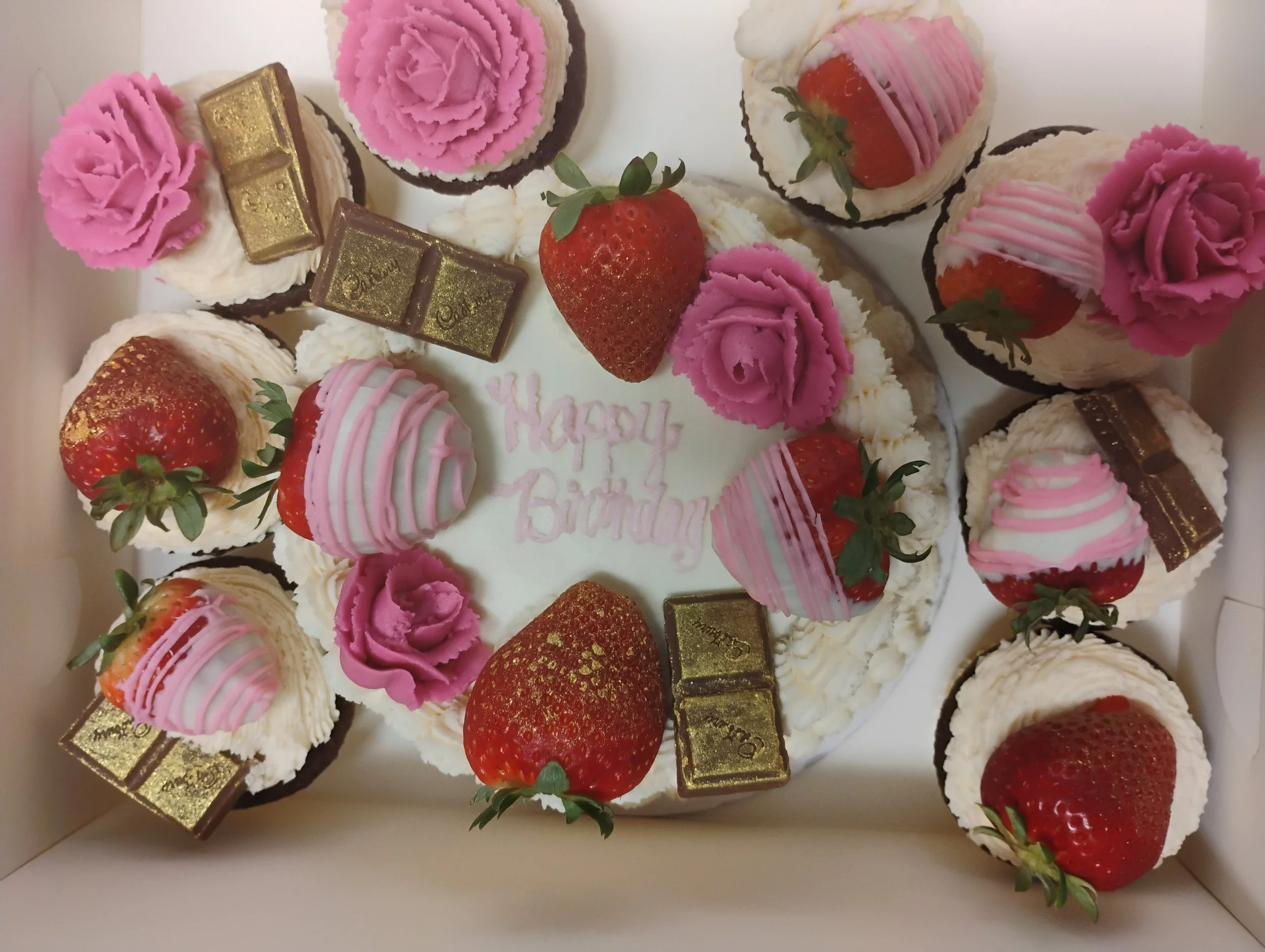 A birthday cake decorated with strawberries, pink roses, gold chocolate pieces, and pink icing, with the message "Happy Birthday" written in pink on the center of the cake.