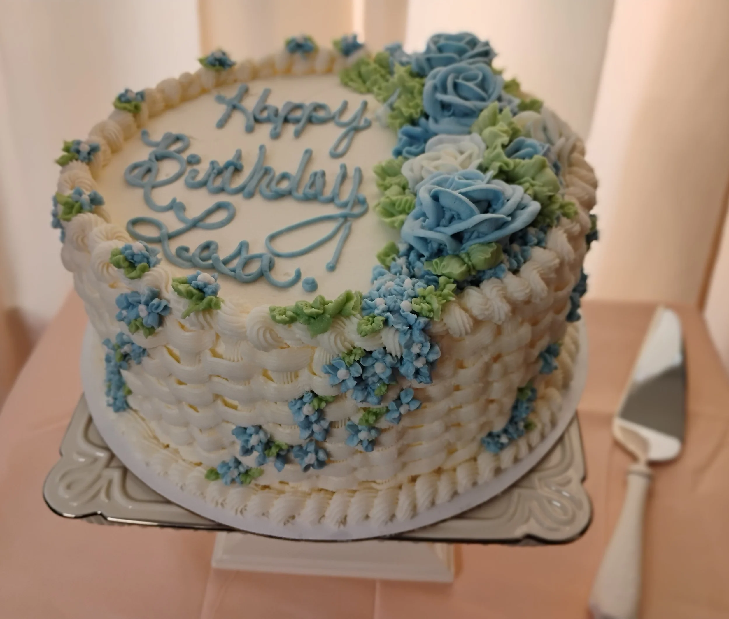 A multi-tiered white birthday cake decorated with blue and green piped roses and small blue flowers, with a birthday message written in blue icing.