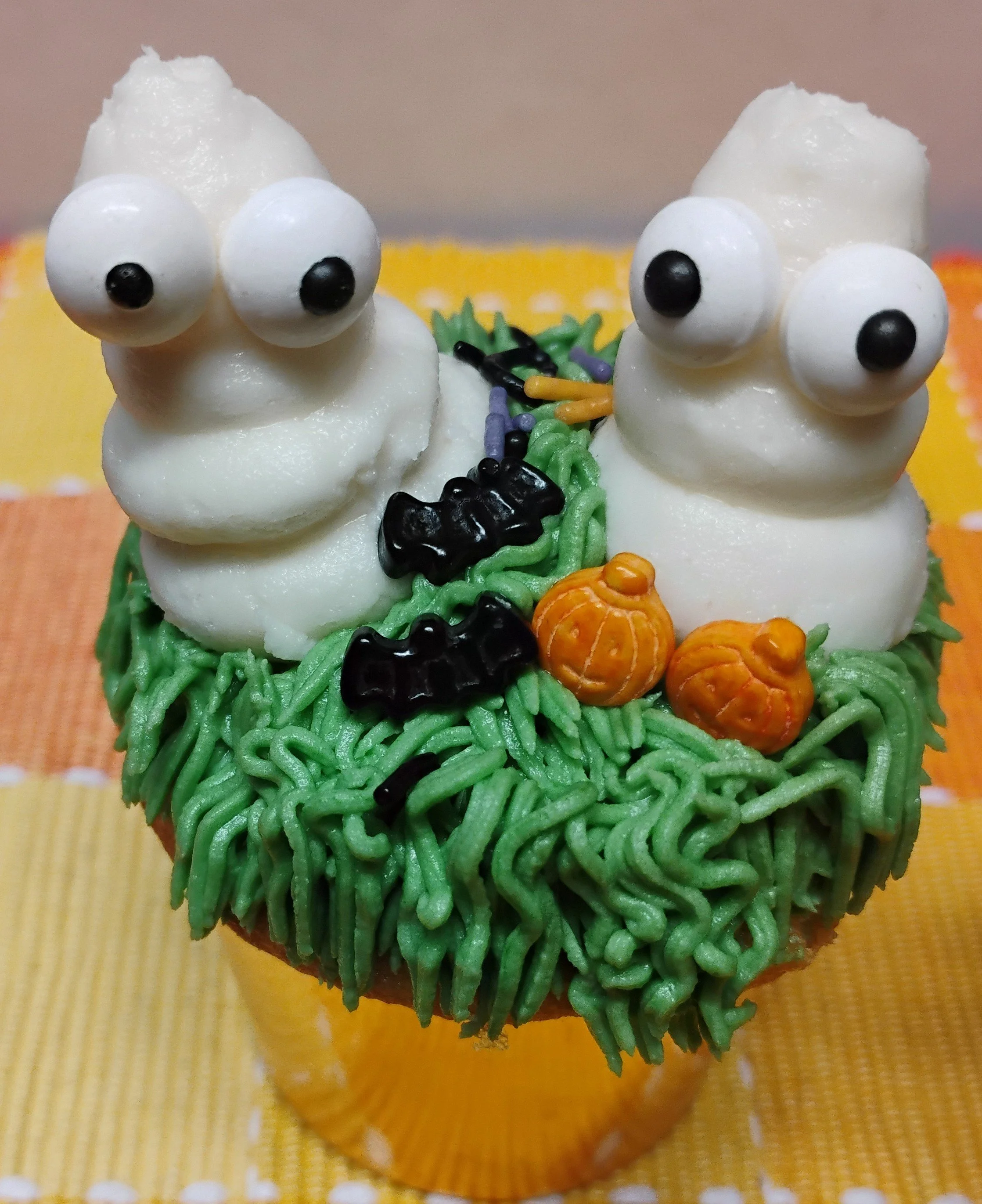 Cupcake decorated with green frosting resembling grass, two white buttercream frosting ghosts with googly eyes, and small Halloween-themed decorations like orange pumpkins and black bats.
