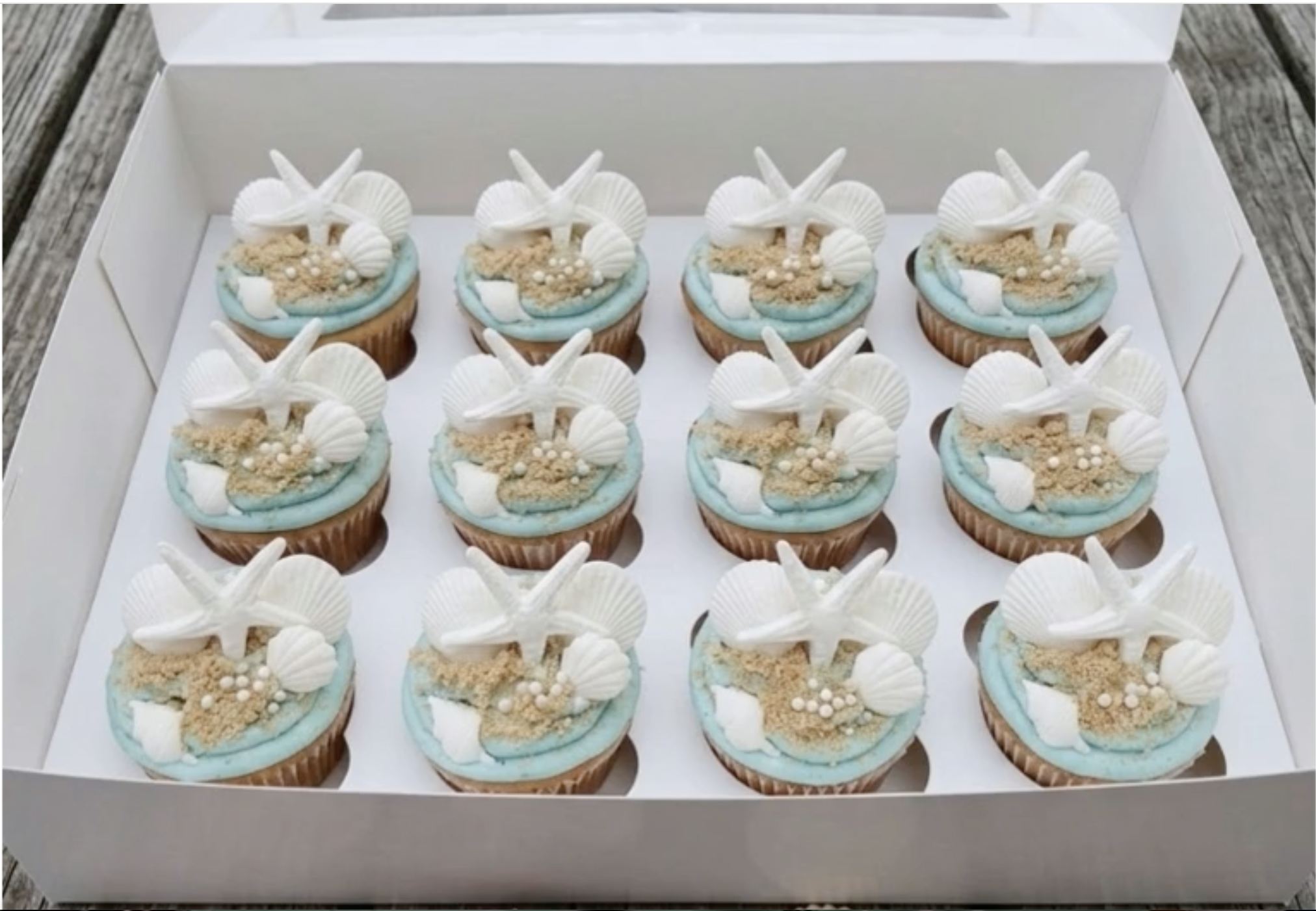A box of nine decorated cupcakes topped with white starfish and shell-shaped decorations, set on a wooden surface.