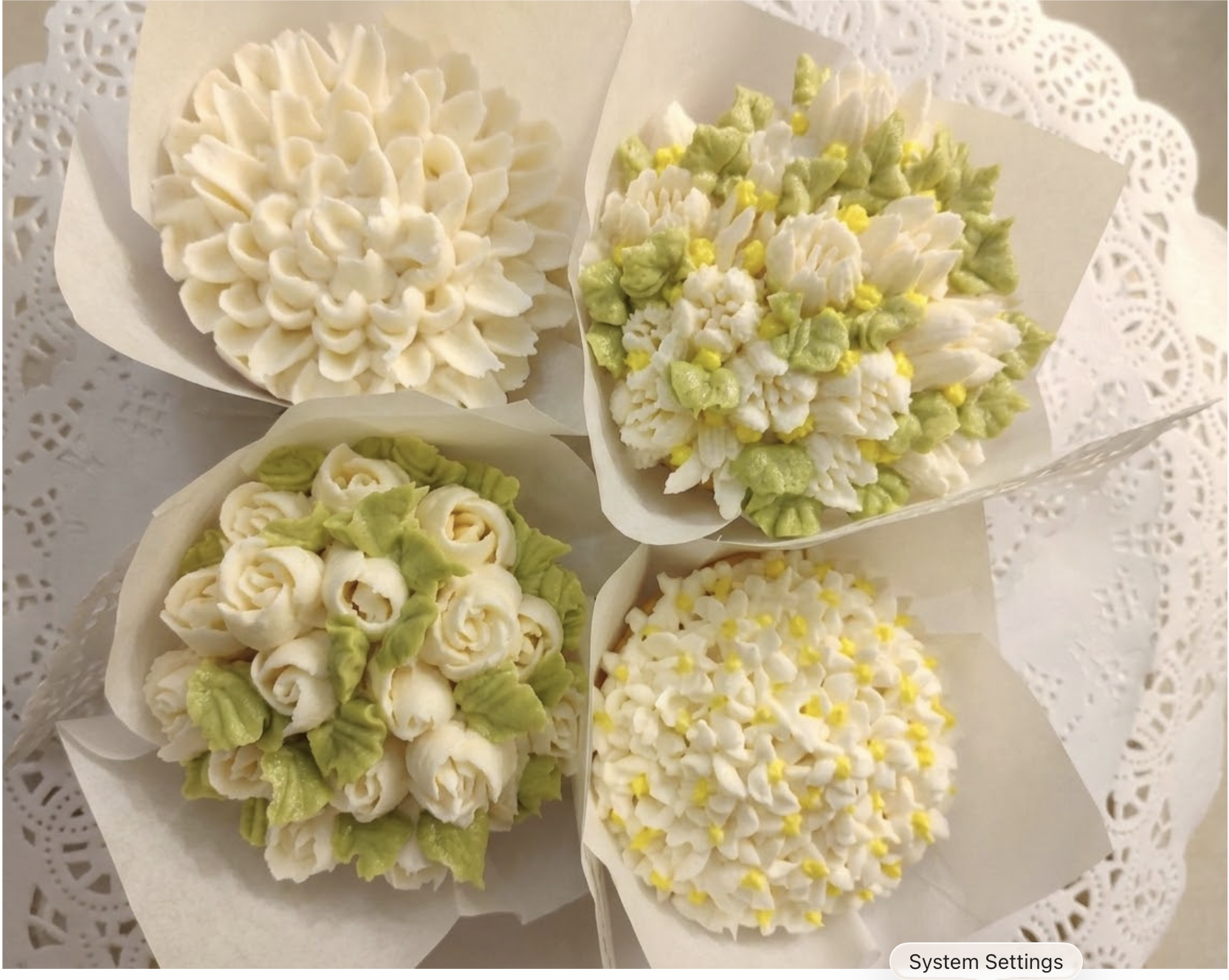 Assorted cupcakes decorated with piped buttercream flowers, some with white, yellow, and green frosting, arranged on a white doily-lined tray.