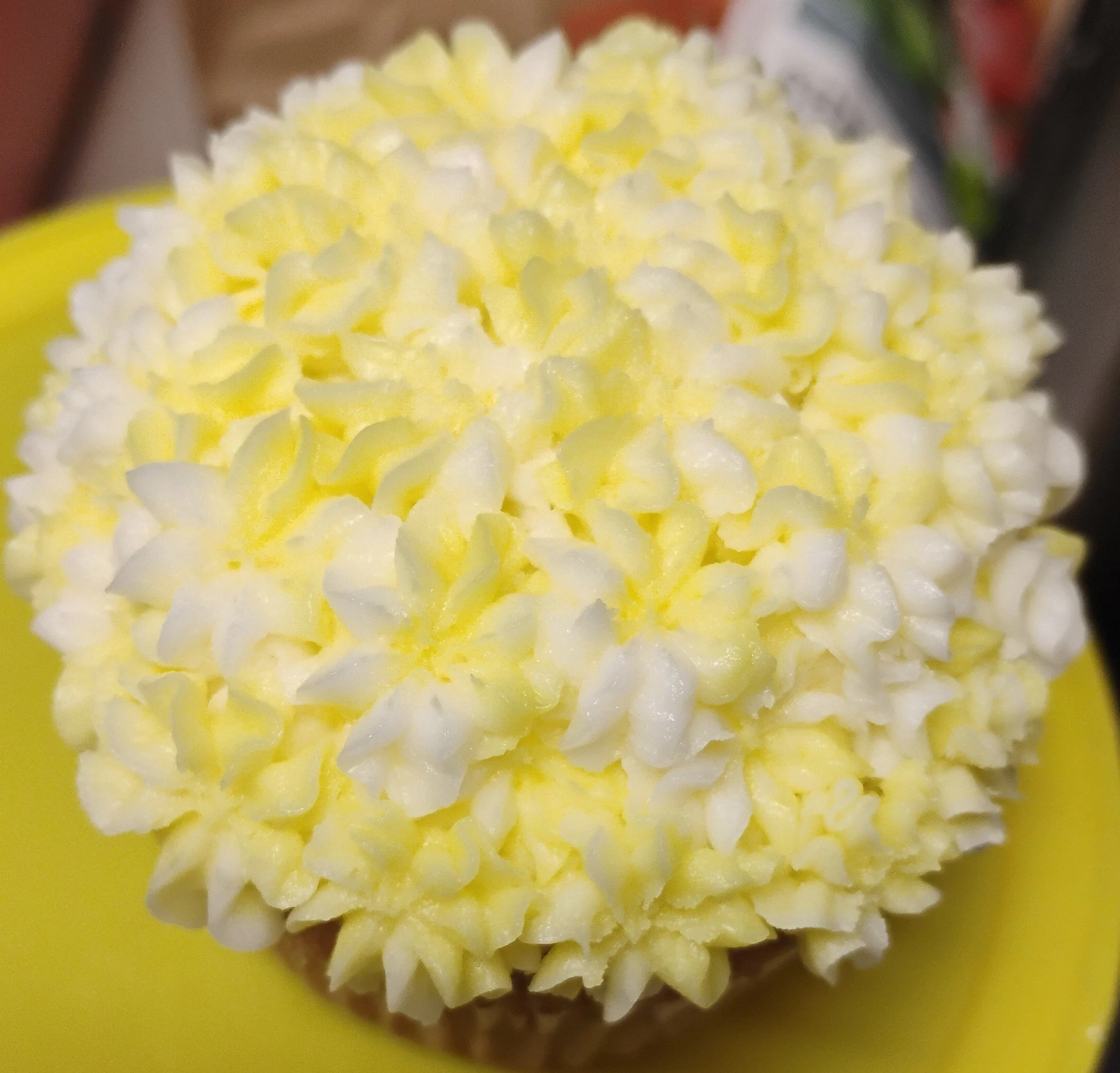 Close-up of a cupcake topped with white and yellow cream or frosting, decorated to look like a flower, on a yellow plate.