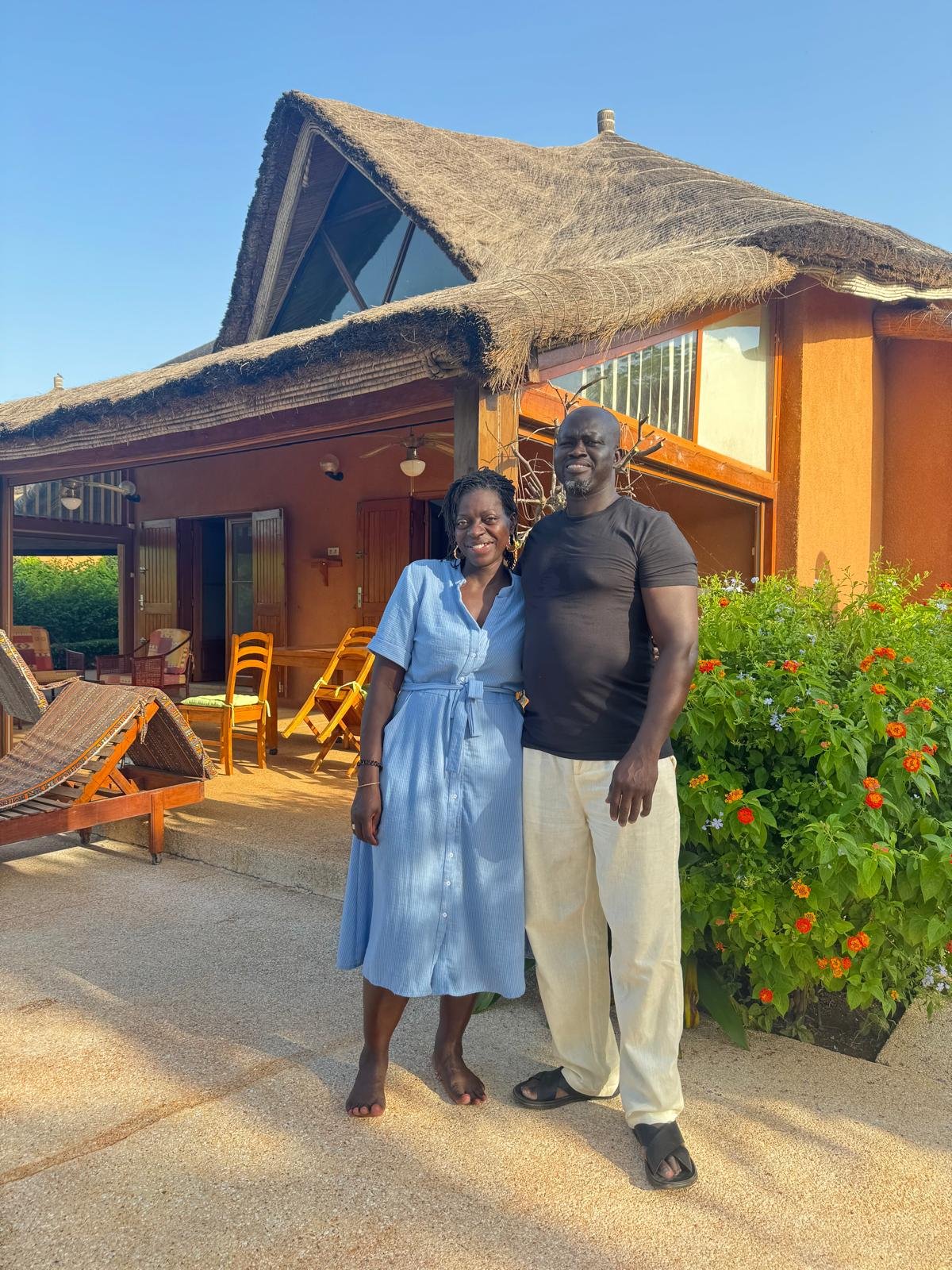 Deux personnes se tiennent devant une maison en style traditionnel africain avec un toit en chaume, sous un ciel bleu, entourées de végétation et de meubles d'extérieur en bois.