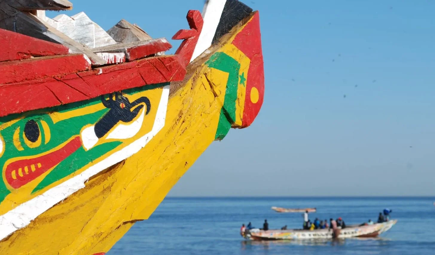Partie avant d'une pirogue colorée avec des motifs africains peints, face à une mer avec un bateau en arrière-plan