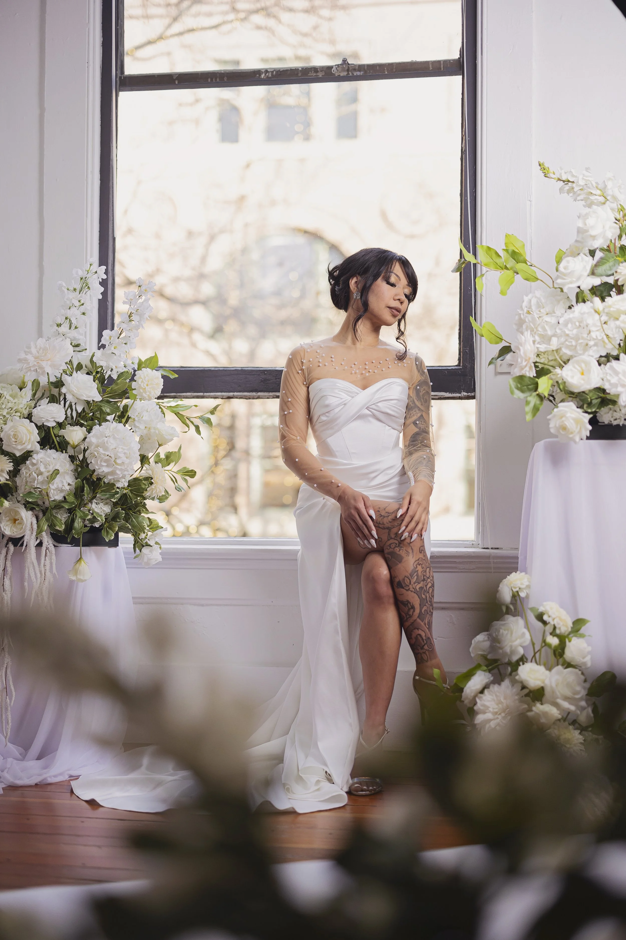 A woman in a white wedding gown sitting by a large window with floral arrangements around her.
