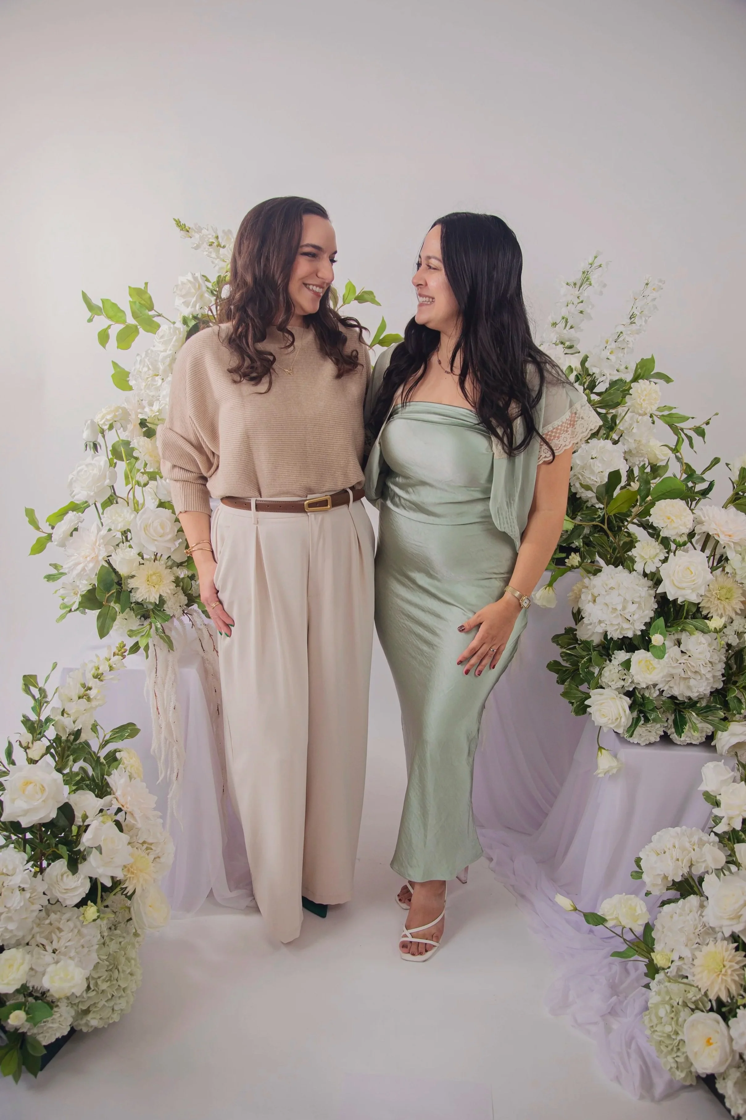 Two women standing close together, smiling, with floral arrangements behind them.
