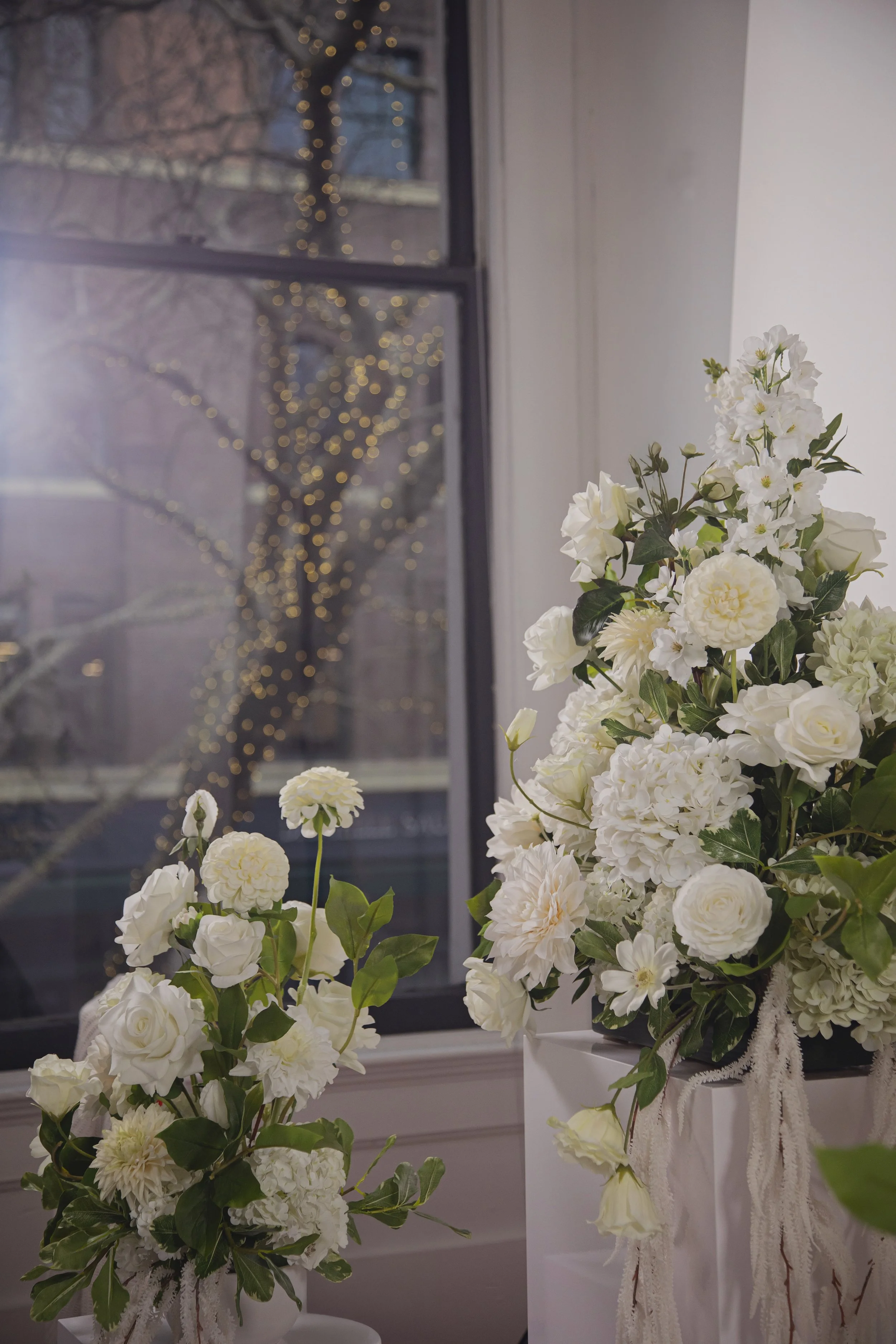 Two large floral arrangements with white flowers and green leaves placed on white stands near a window with blurry tree and lights outside.