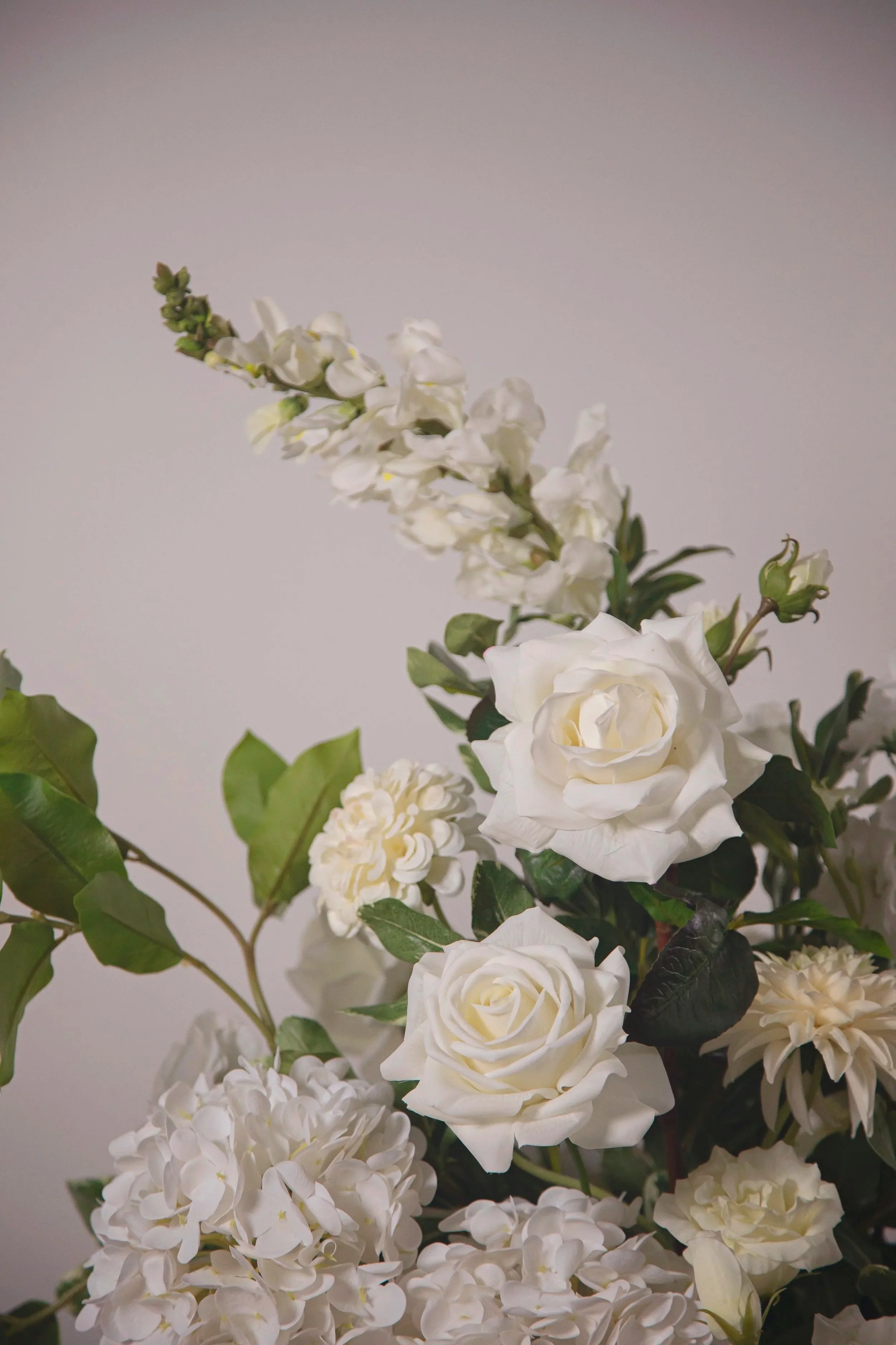 A bouquet of white roses, hydrangeas, and other flowers with green leaves against a plain light background.