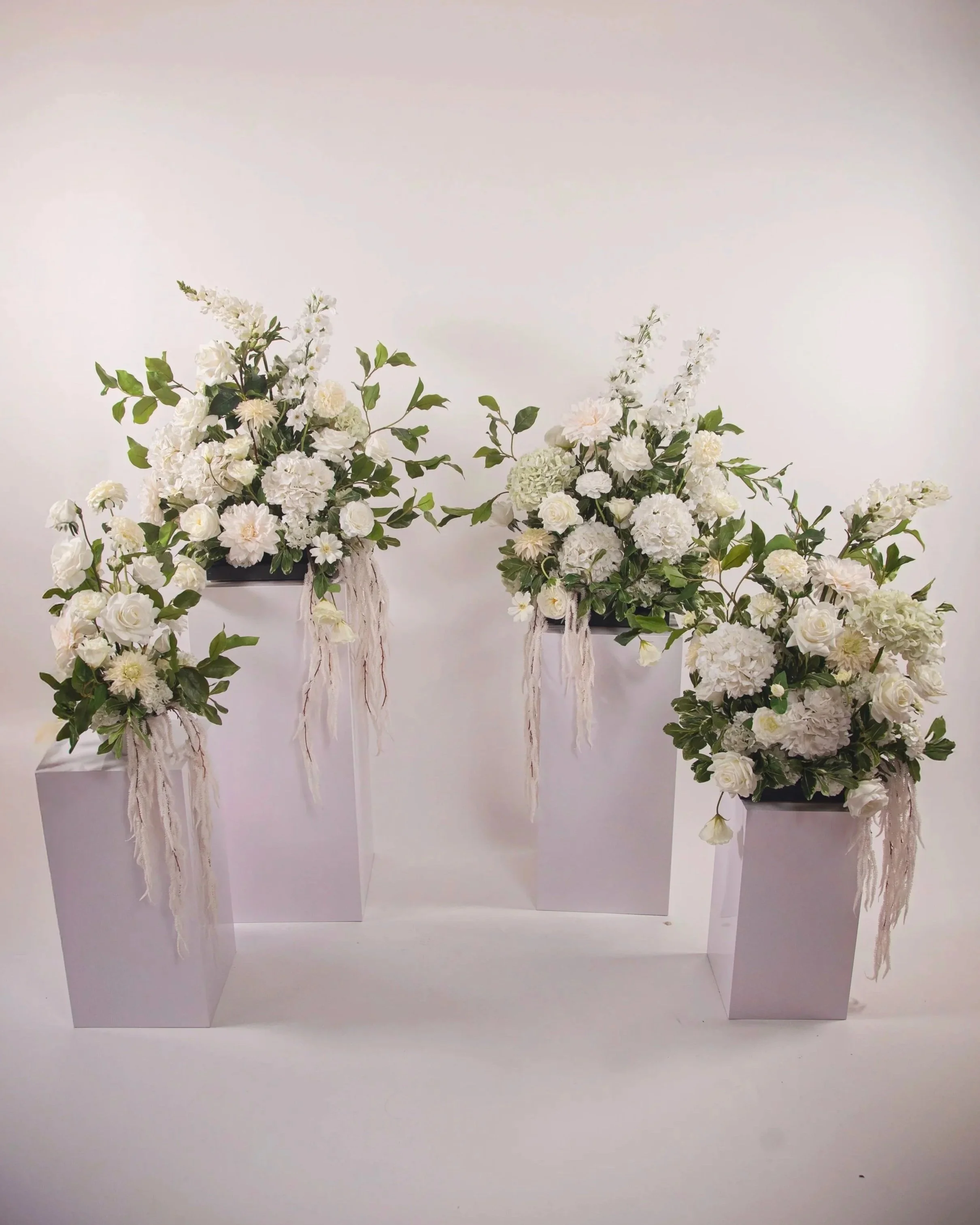 Three white floral arrangements in white square vases on white pedestals against a white background.