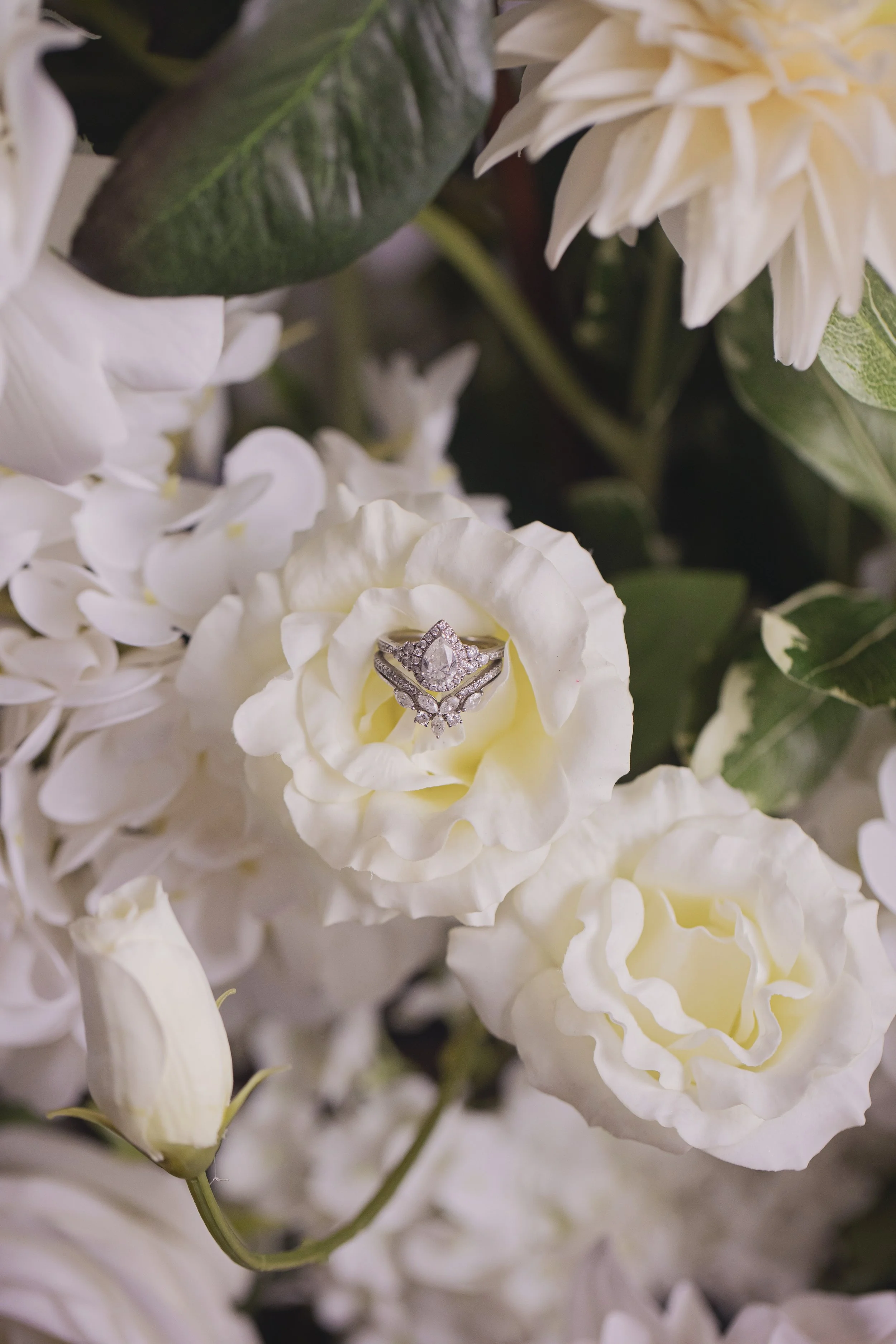 Close-up of a diamond engagement ring placed on white flowers, including roses and hydrangeas, with green leaves in the background.