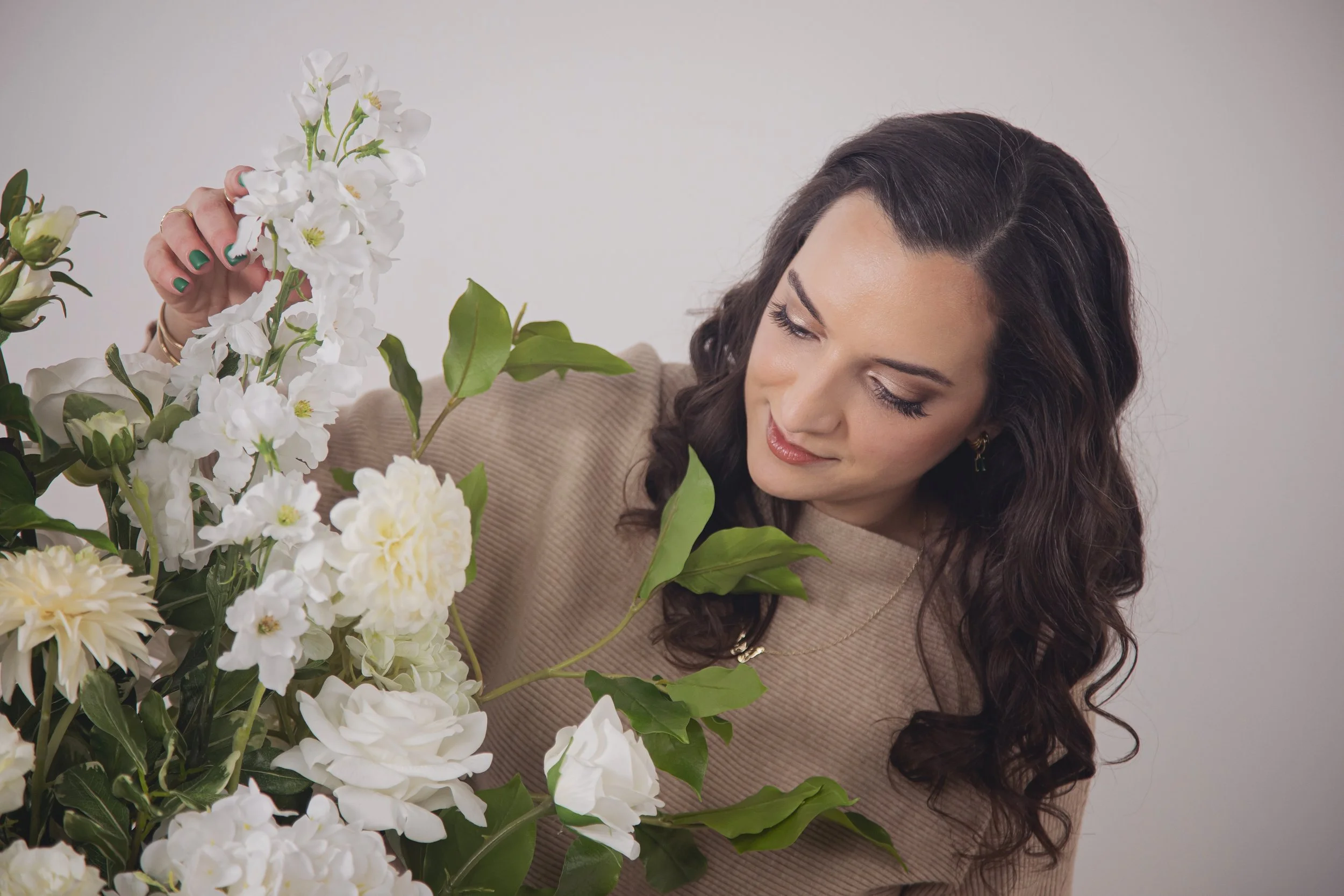 A woman with long, wavy brown hair wearing a beige sweater arranging white and cream-colored flowers in a vase.
