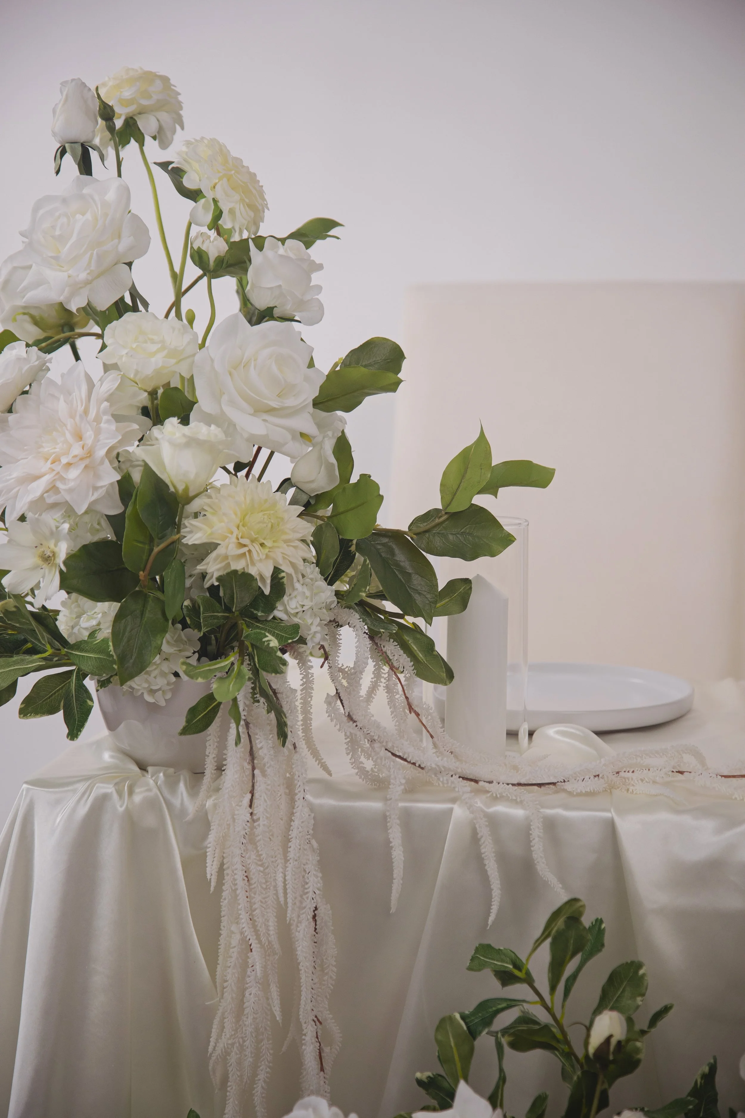 A white floral arrangement with roses and other flowers on a satin tablecloth, with white candles and plates in the background.
