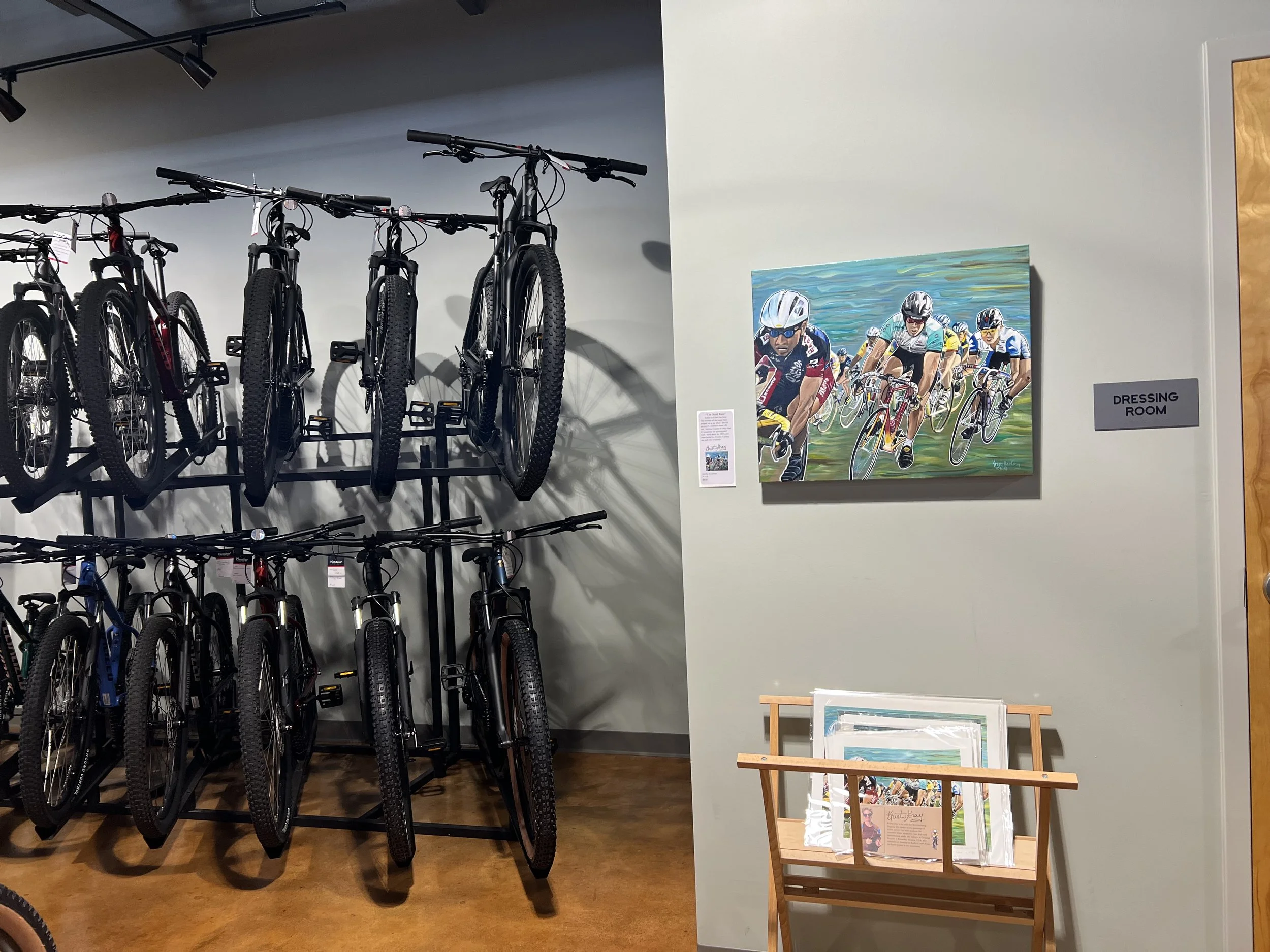 The Good Race original acrylic painting of a 1990 criterium race in Richmond, Virginia at 20 inches by 24 inches next to a rack of bicycles and over a rack of quality art prints in Cardinal Bicycle in Grandin Village, Roanoke, Virginia.