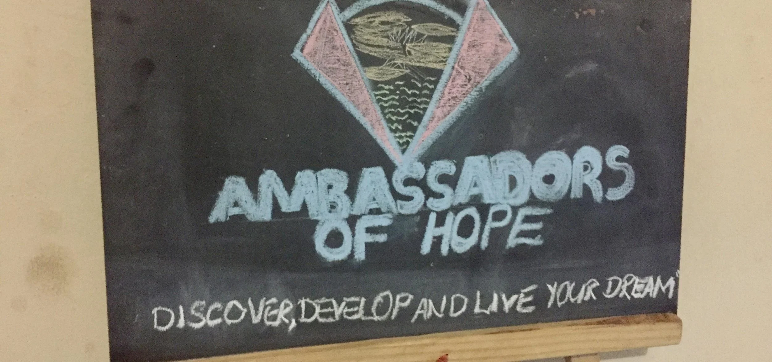 Chalkboard with a colorful triangle logo above the words "Ambassadors of Hope." Below, a phrase reads "Discover, Develop and Live Your Dream."