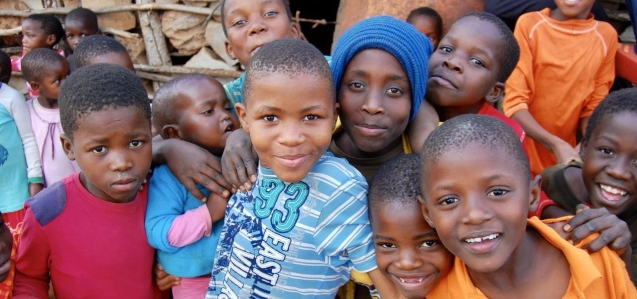 A group of diverse children smiling and posing outdoors, with some hugging and others looking at the camera, in front of a rustic background.