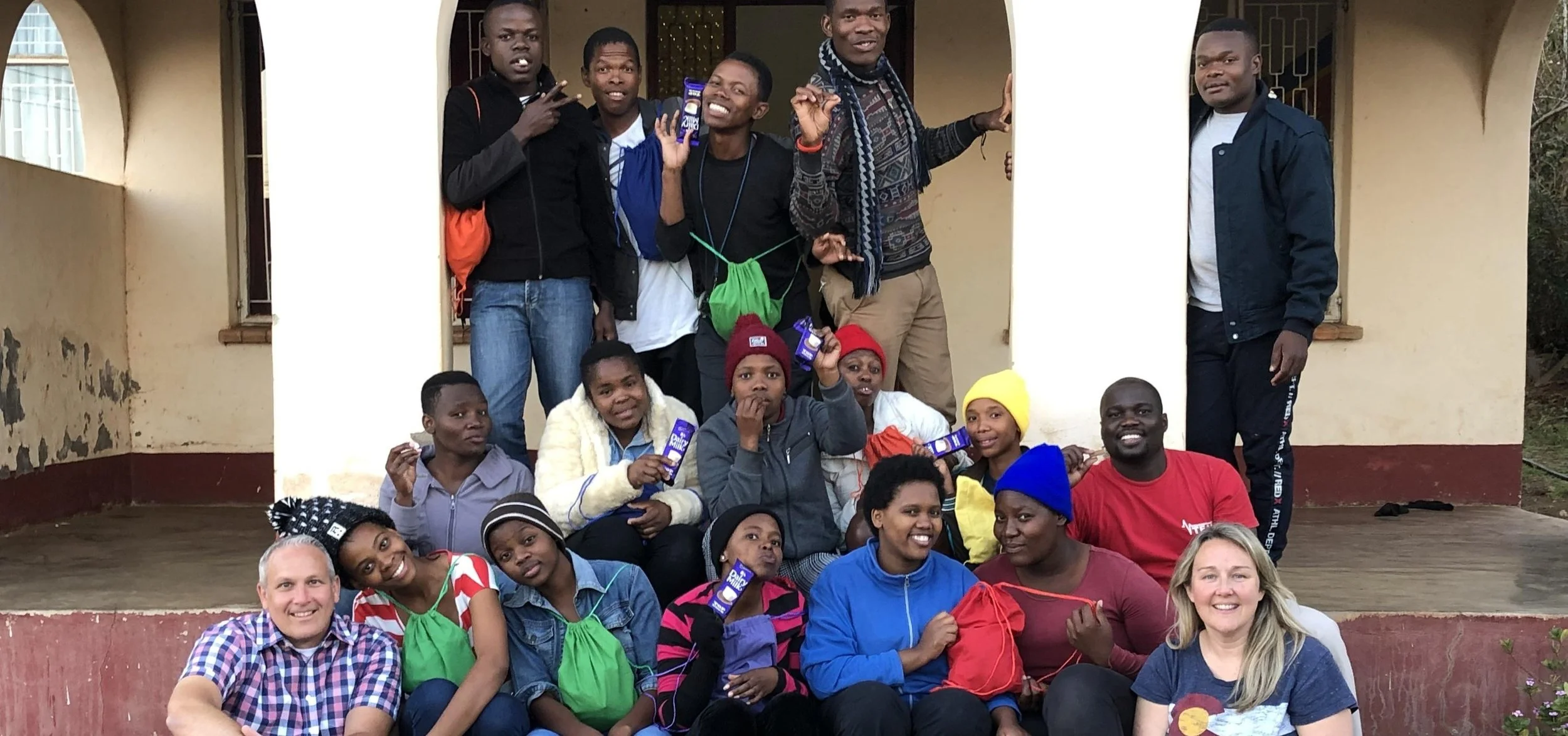 Group of diverse young people and two adults posing outside a building, some holding chocolates, smiling and making peace signs.