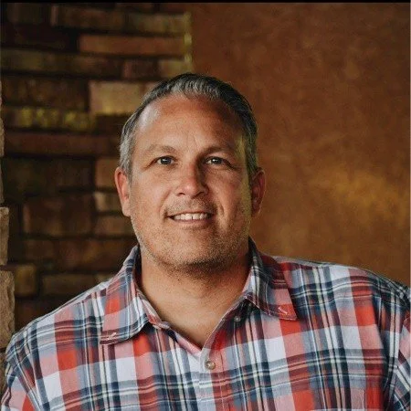 Man with short gray hair wearing a red, blue, and white plaid shirt smiling indoors with brick and brown walls in the background.