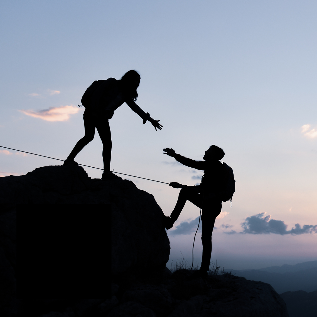 Silhouette of a woman helping a man climb a mountain during sunset.