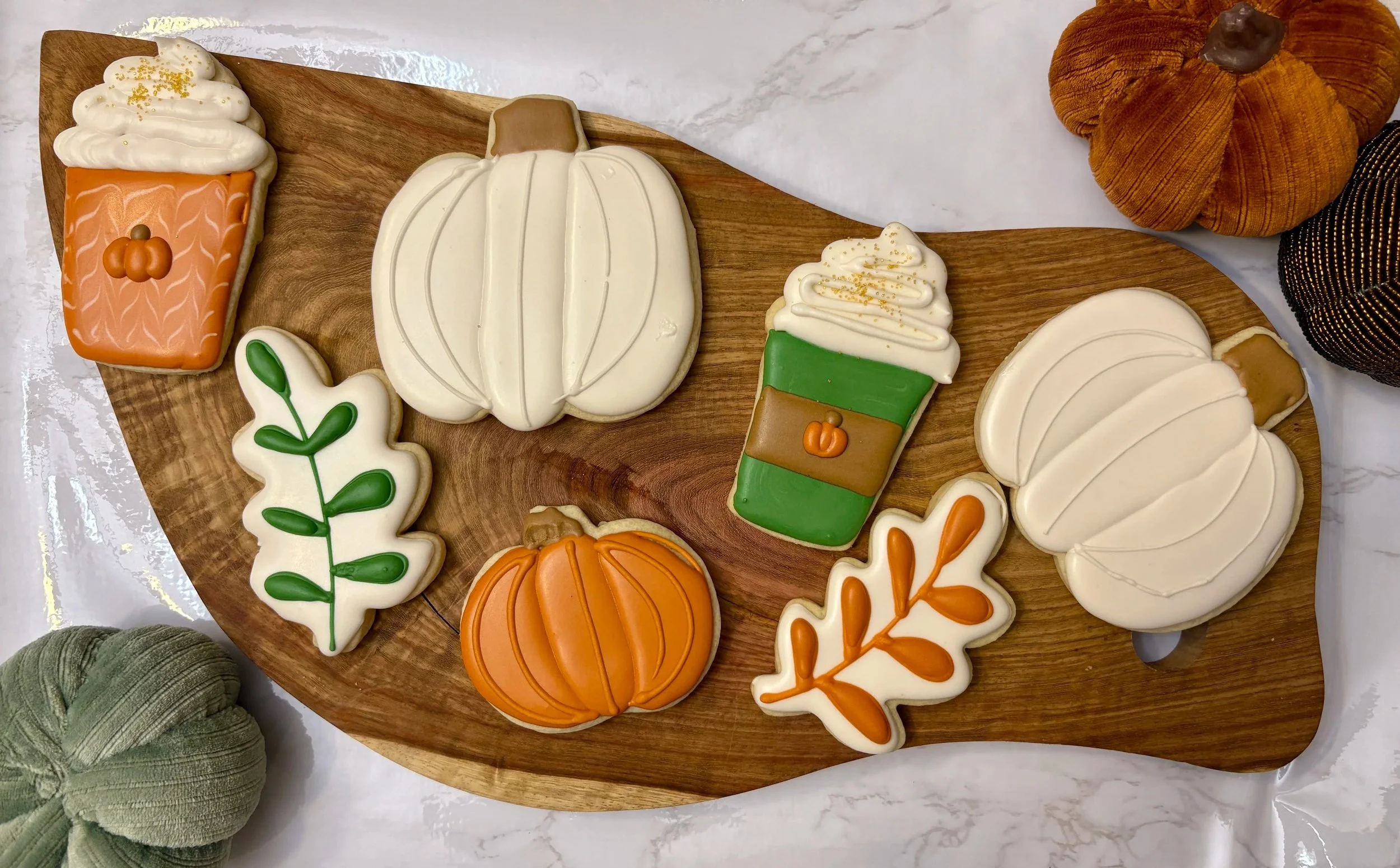 Assorted pumpkin-shaped and autumn-themed cookies decorated with colorful icing, arranged on a wooden platter. Includes white, orange, and green decorated cookies with leaf and pumpkin motifs.