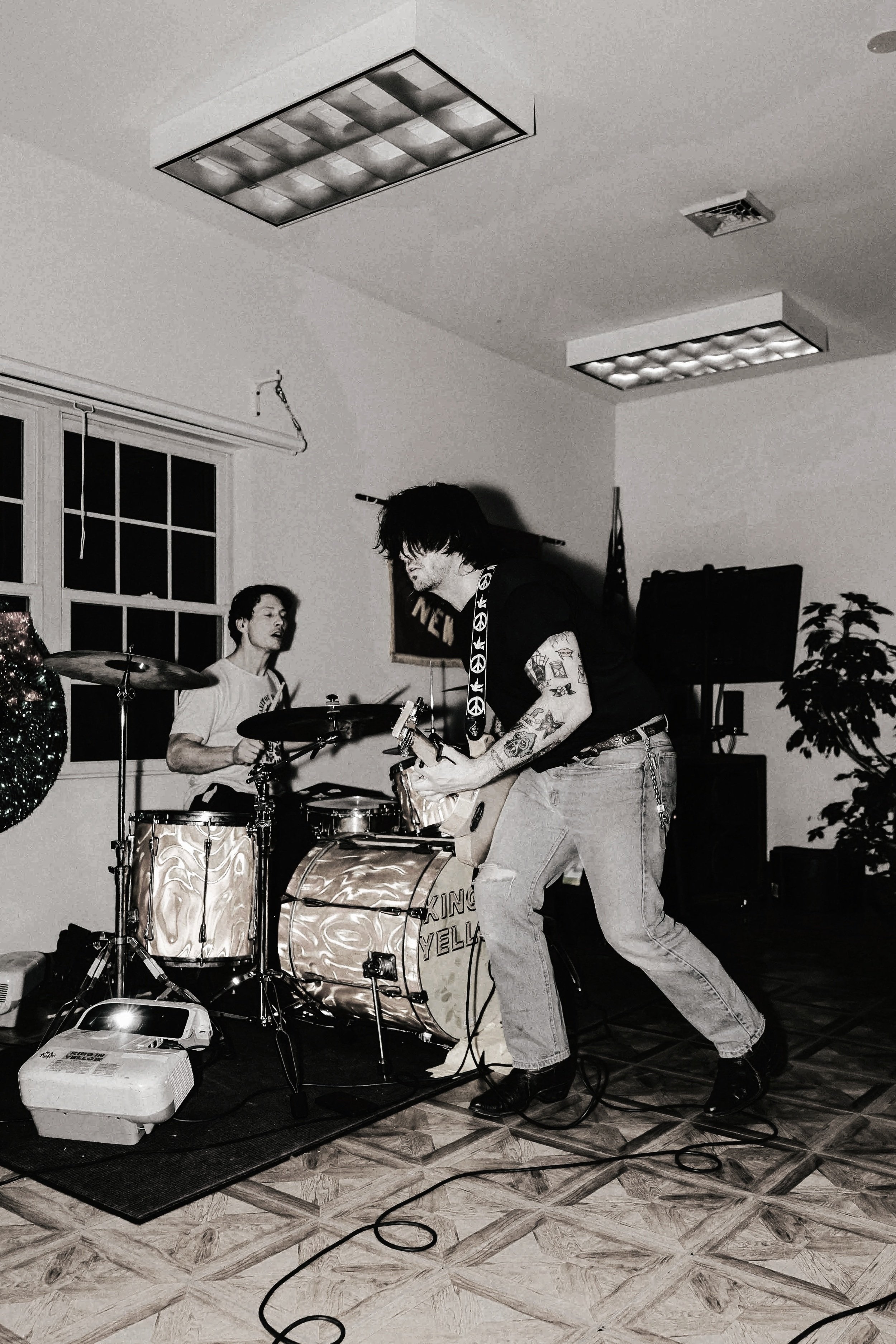 Black and white photo of a musician playing a drum set and a man playing a keyboard in a room with tiled floor, window, and ceiling lights.