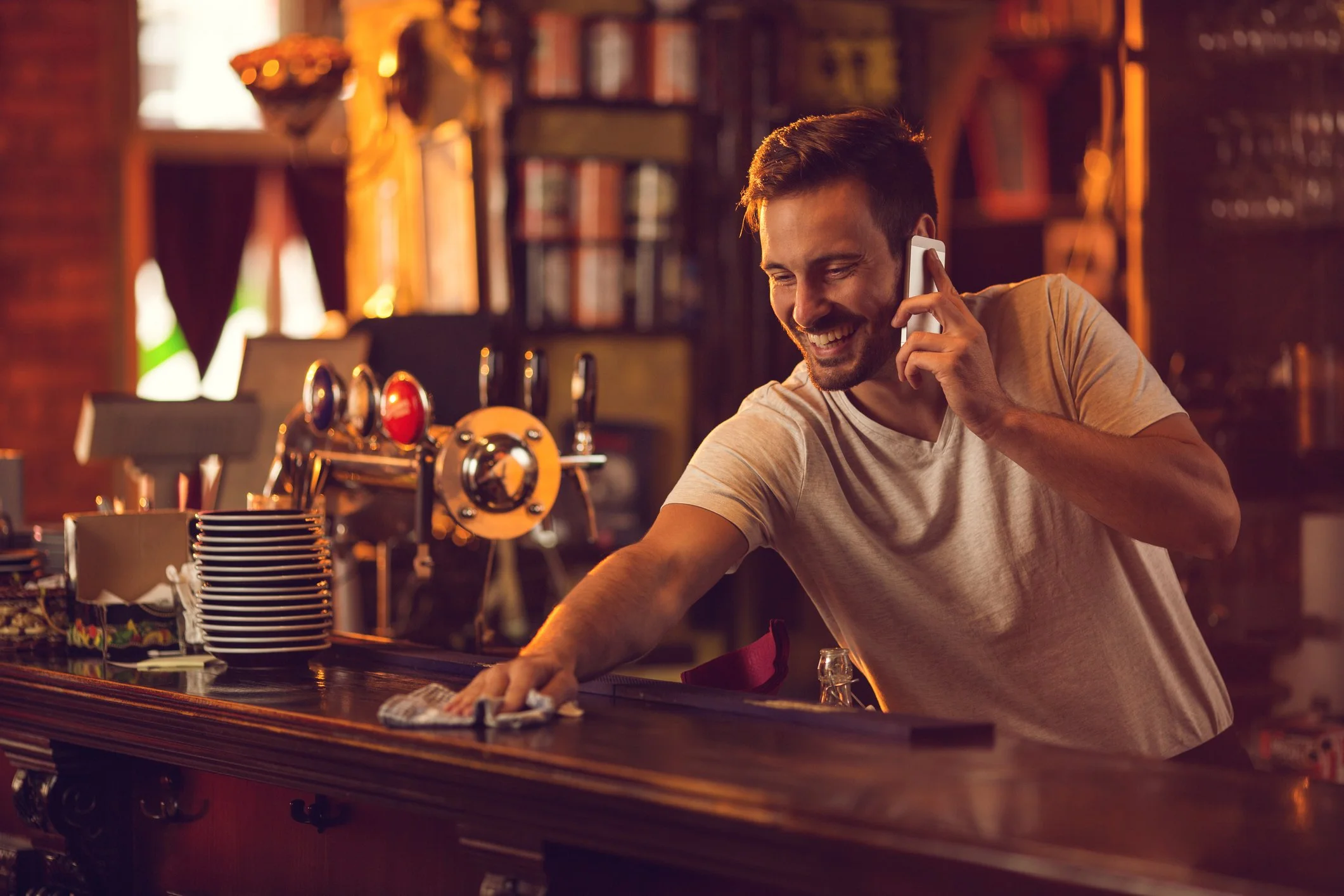 A smiling bartender cleaning the bar counter in a dimly lit bar with beer tap handles in the background.