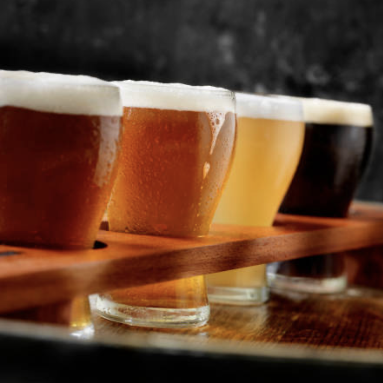 Four glasses of beer on a wooden paddle, with different colors from dark to light, against a dark background.