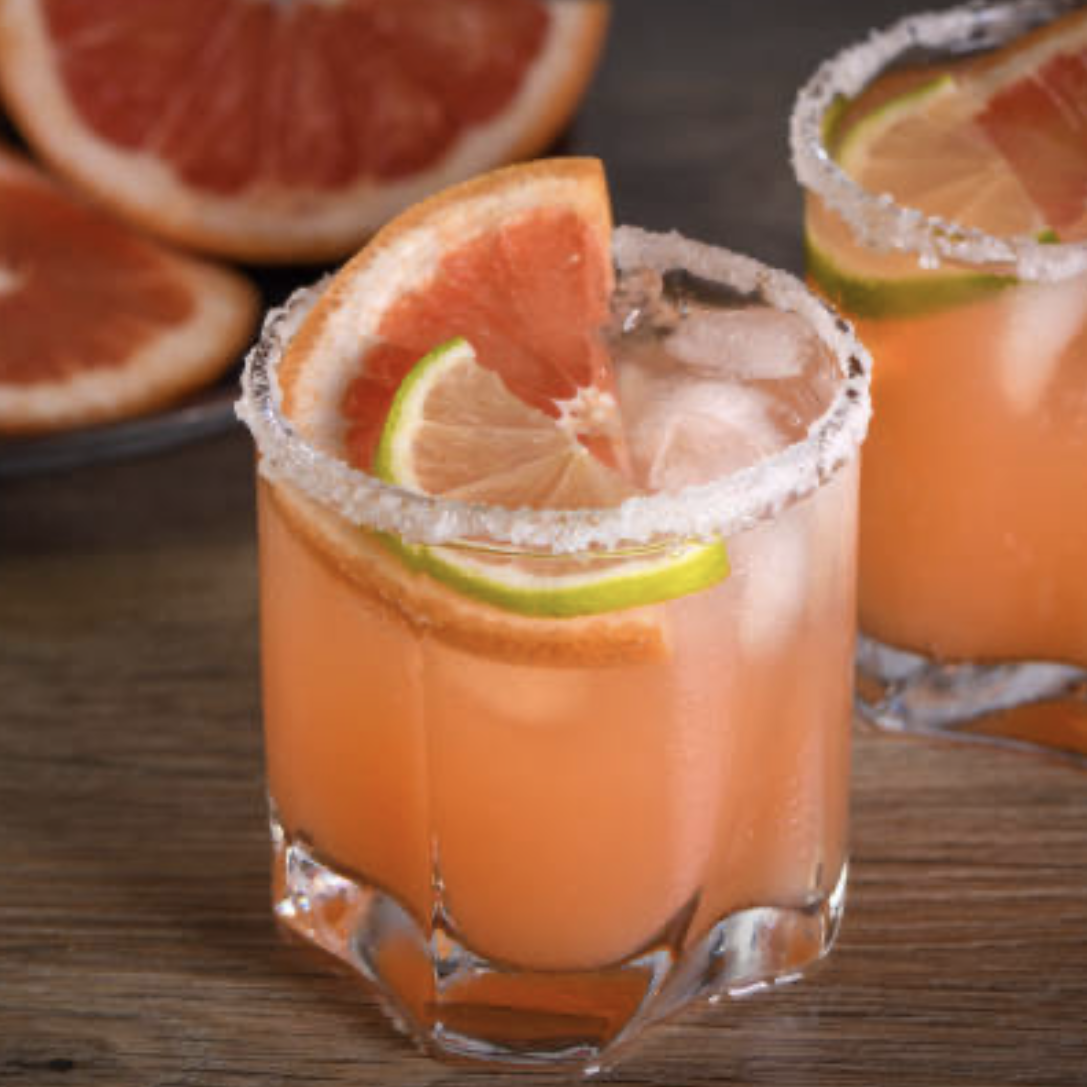 Two glasses of grapefruit cocktails with lime and grapefruit slices, salted rims, and ice, on a wooden surface with grapefruit halves in the background.